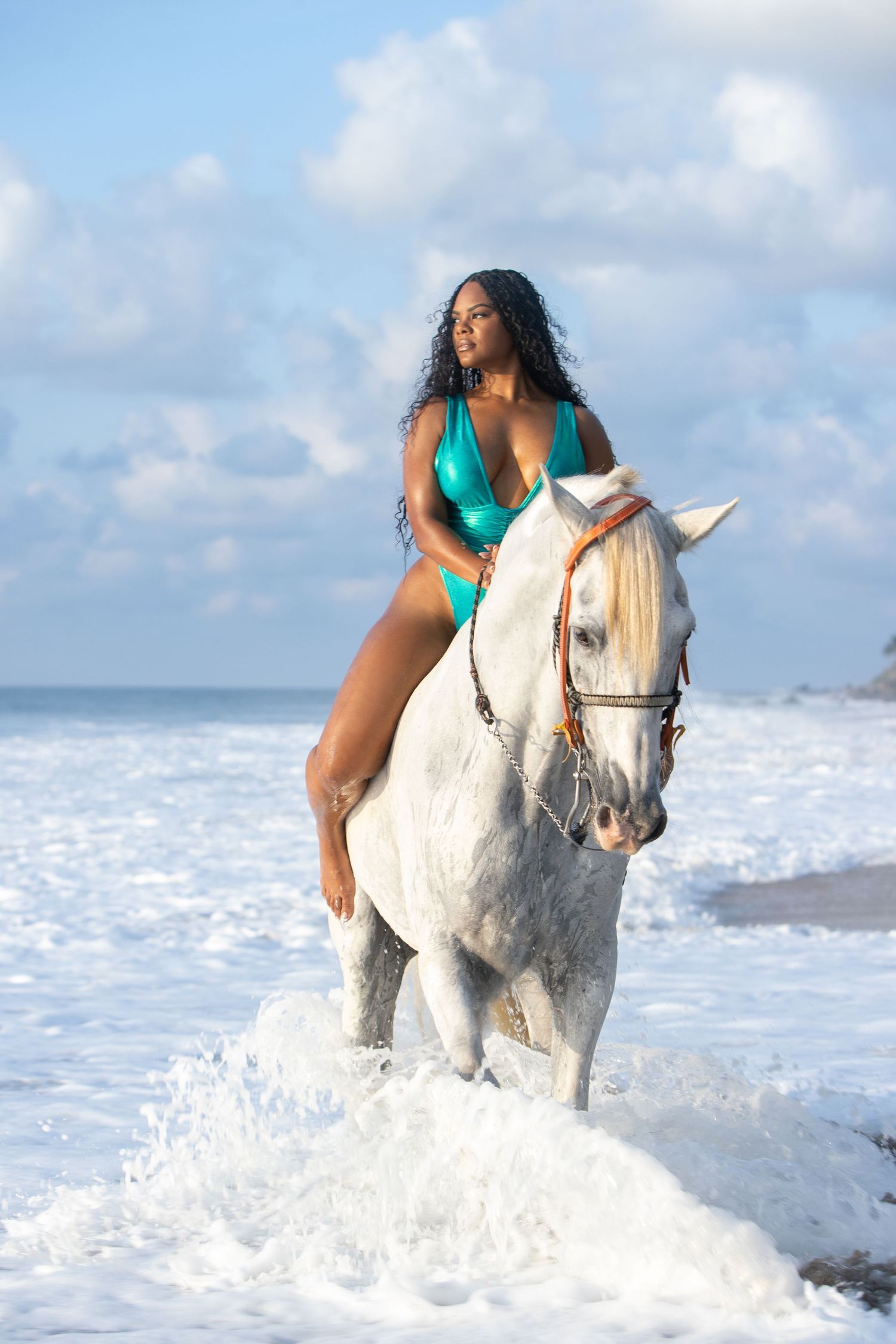 A rider in a turquoise swimsuit guides a white horse through crashing ocean waves on a sunny beach.