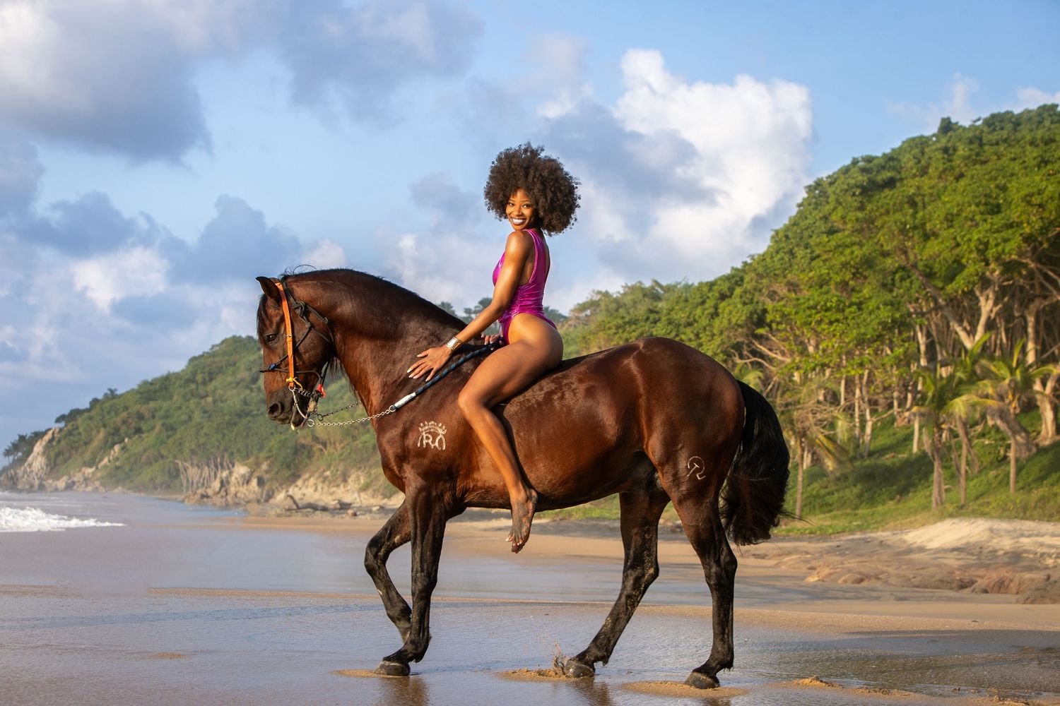 A rider on horseback walking along a tropical beach with lush green hills in the background.