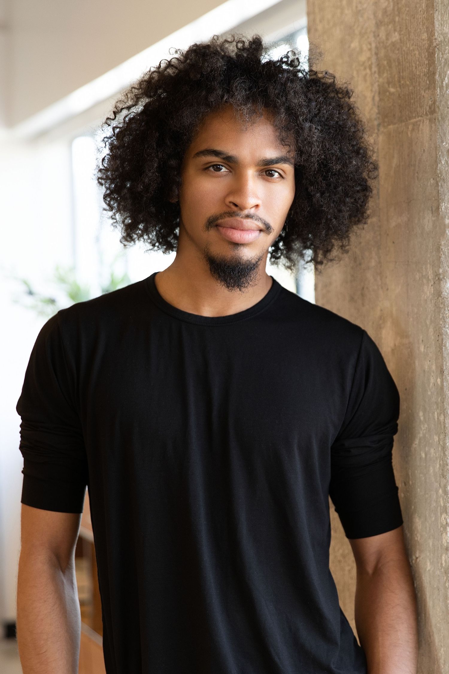 Person with natural curly afro hairstyle wearing a black t-shirt poses against a beige wall in natural lighting.