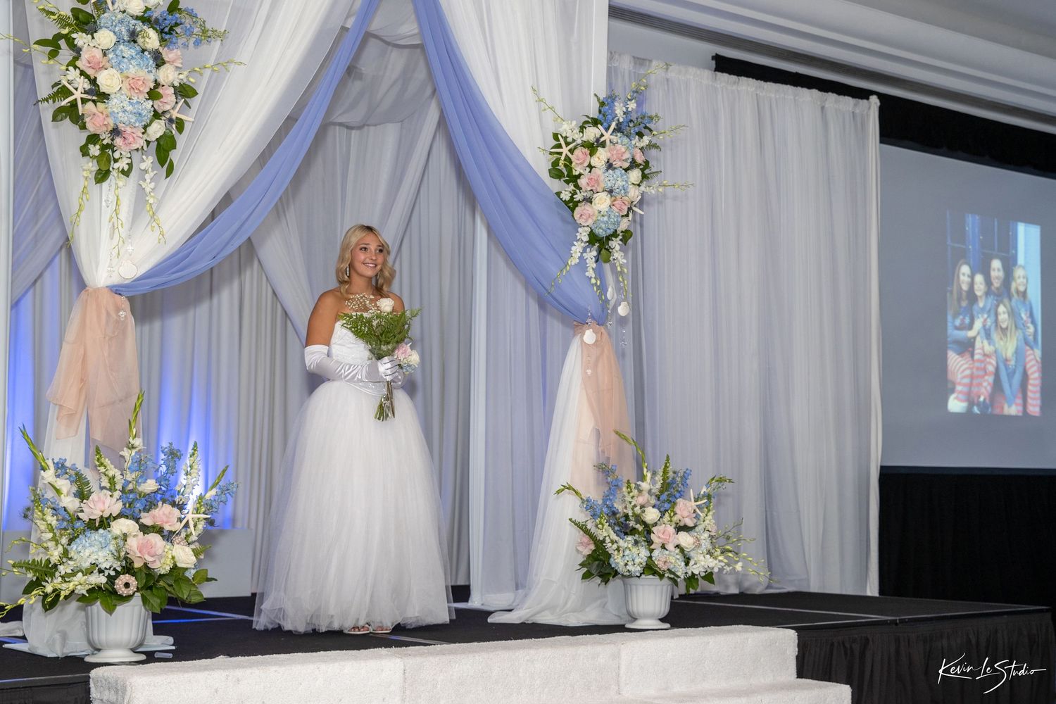 Elegant white wedding reception hall with draped fabric, floral arrangements, and blue accent lighting along the walls.
