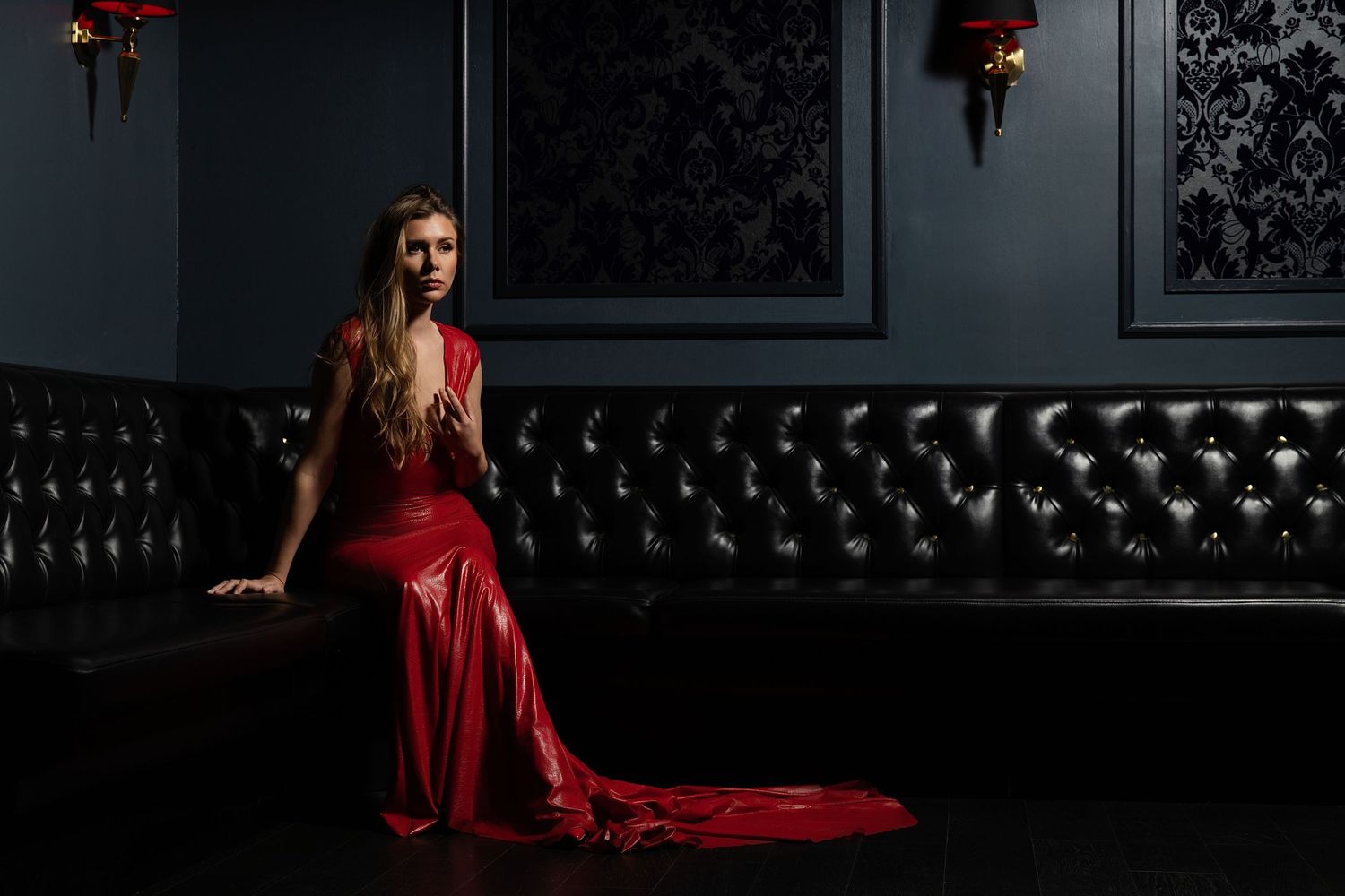 A dramatic fashion photo of someone in a flowing red gown sitting on a black leather couch in a dark moody setting.