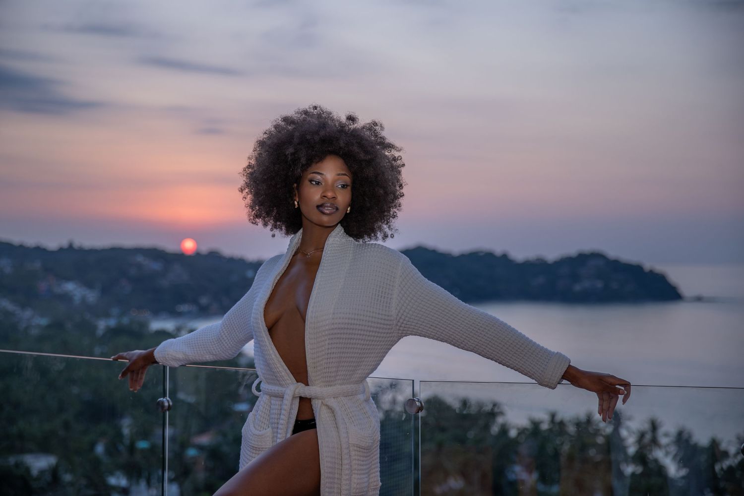 Woman in white dress poses against dramatic coastal sunset backdrop with ocean and mountains in view.