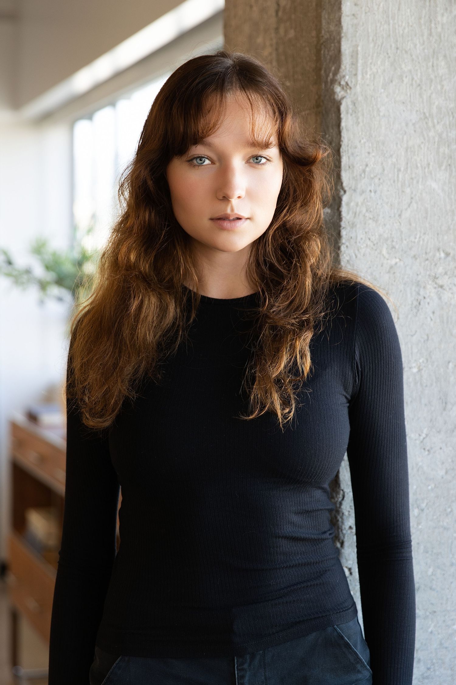 Person wearing a black long-sleeved top stands against a light-colored wall with soft natural lighting.