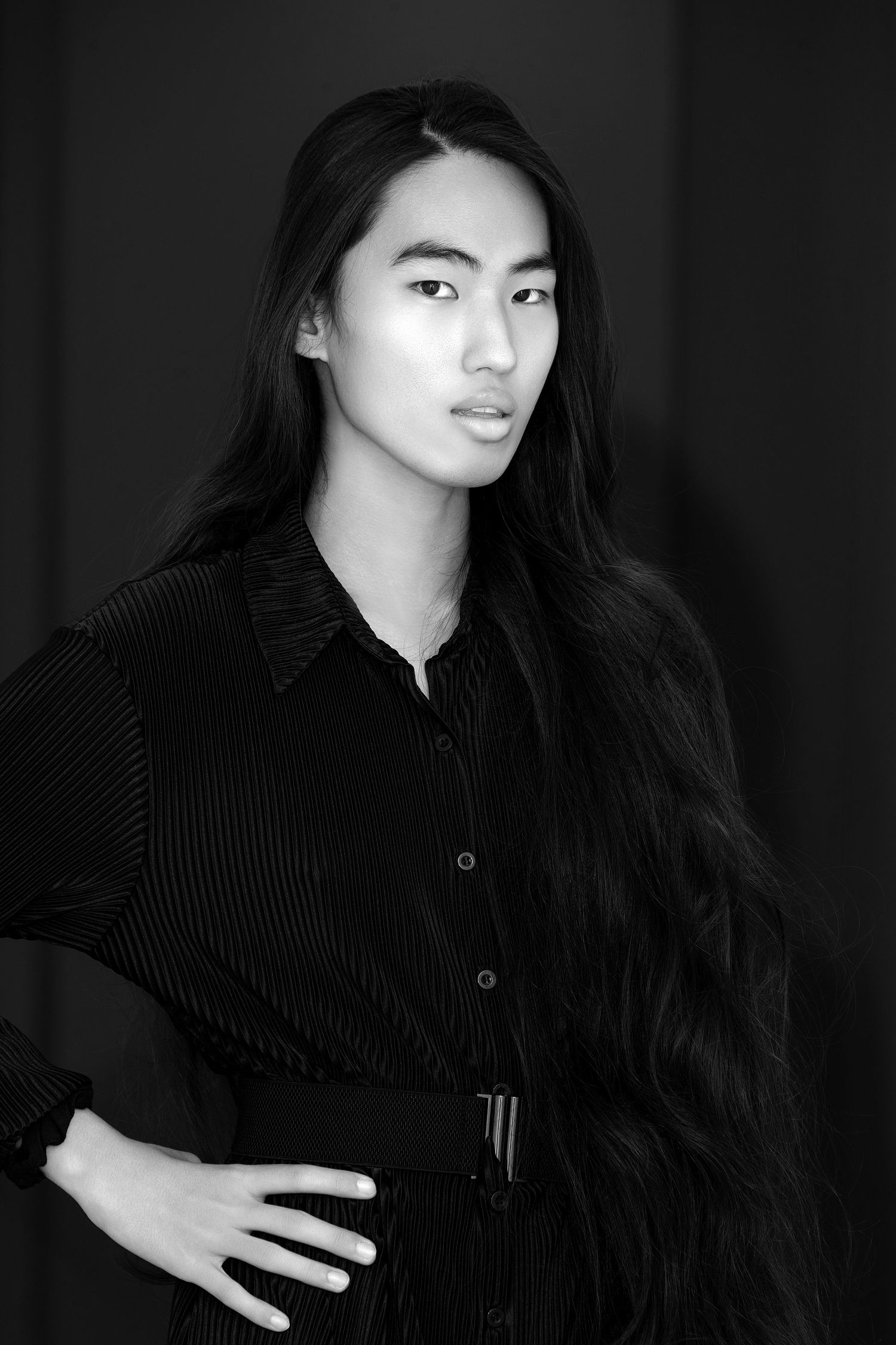 Black and white portrait of a person with long dark hair wearing a black button-up top against a dark background.