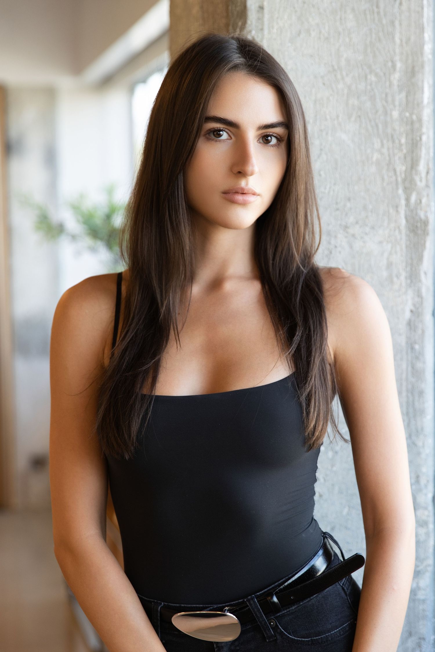 A woman in a black tank top and jeans poses against a light wall in an indoor setting.