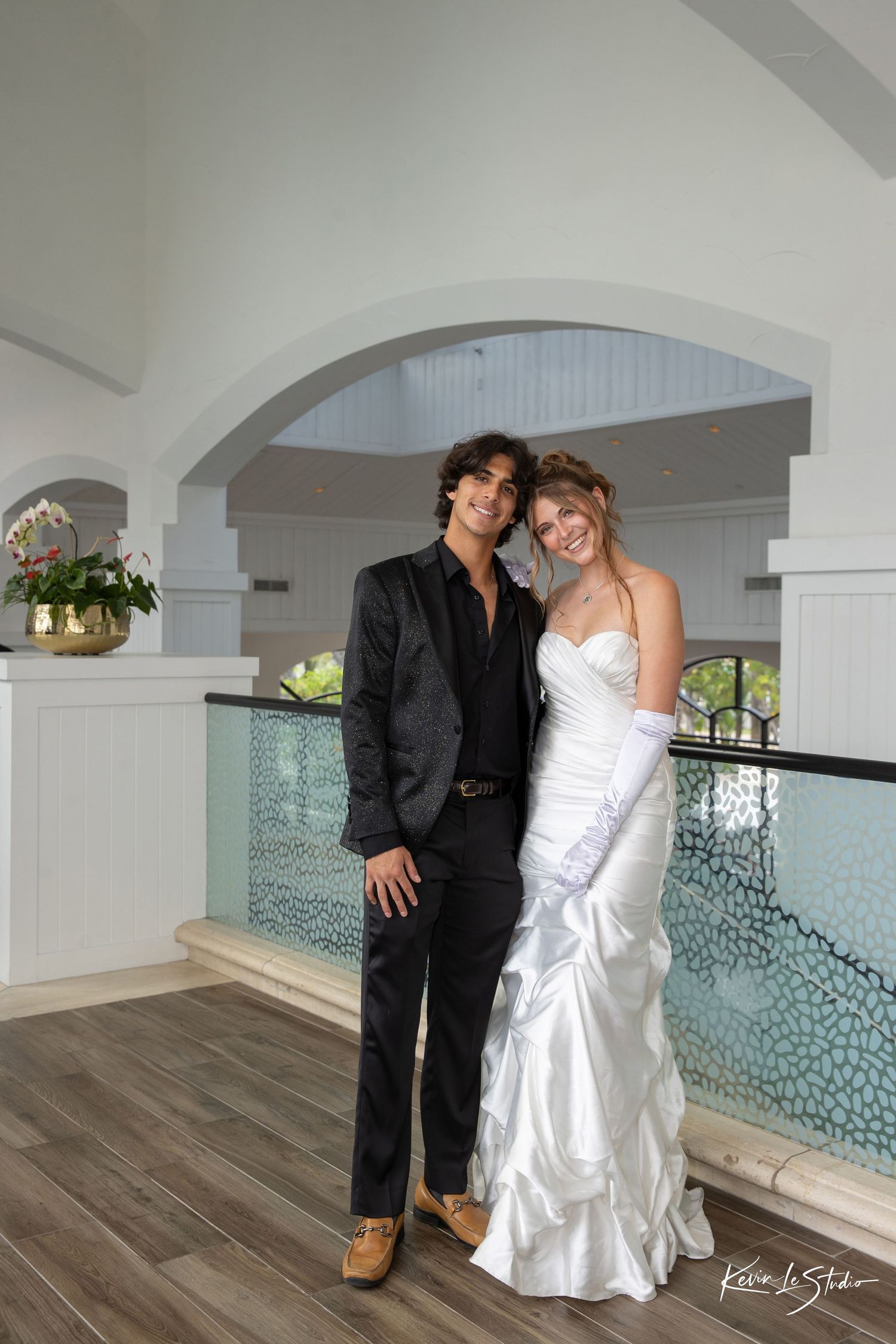 A couple poses together at an elegant venue with arched doorways and glass railings, the woman in white and man in black attire.