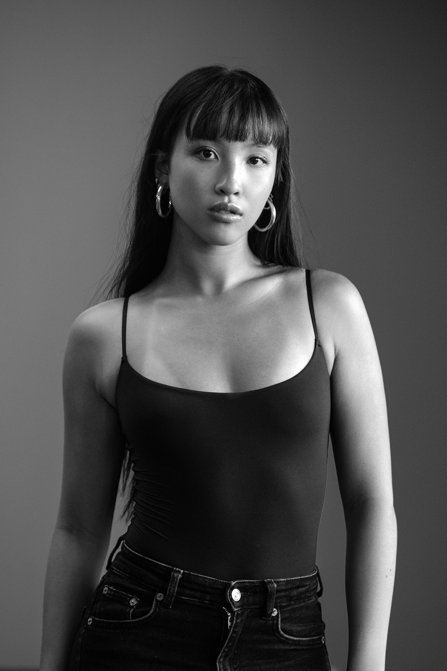Black and white portrait of someone wearing a black tank top and jeans against a dark background.