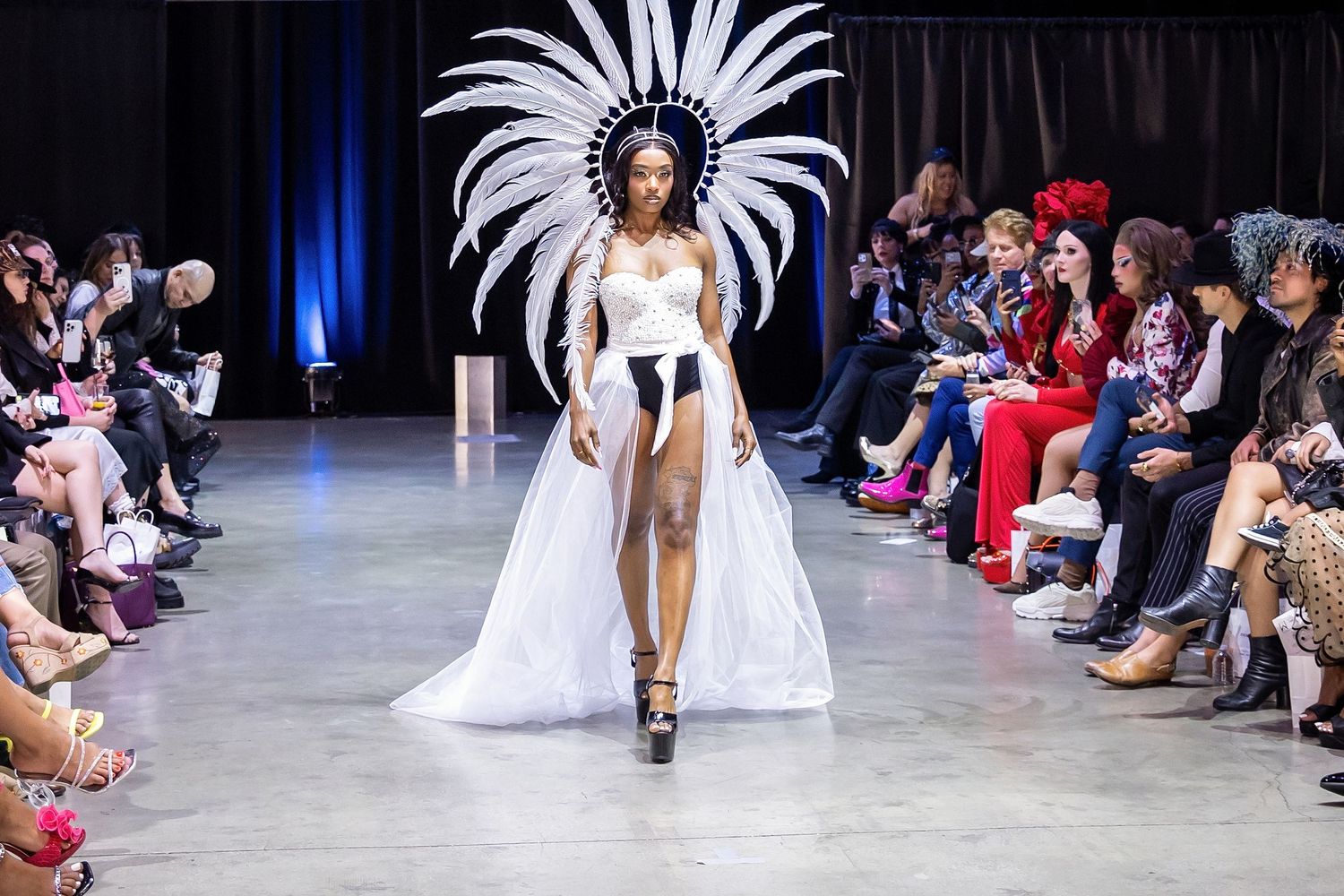 A model walks the runway in a dramatic white gown with a feathered headdress during a fashion show.
