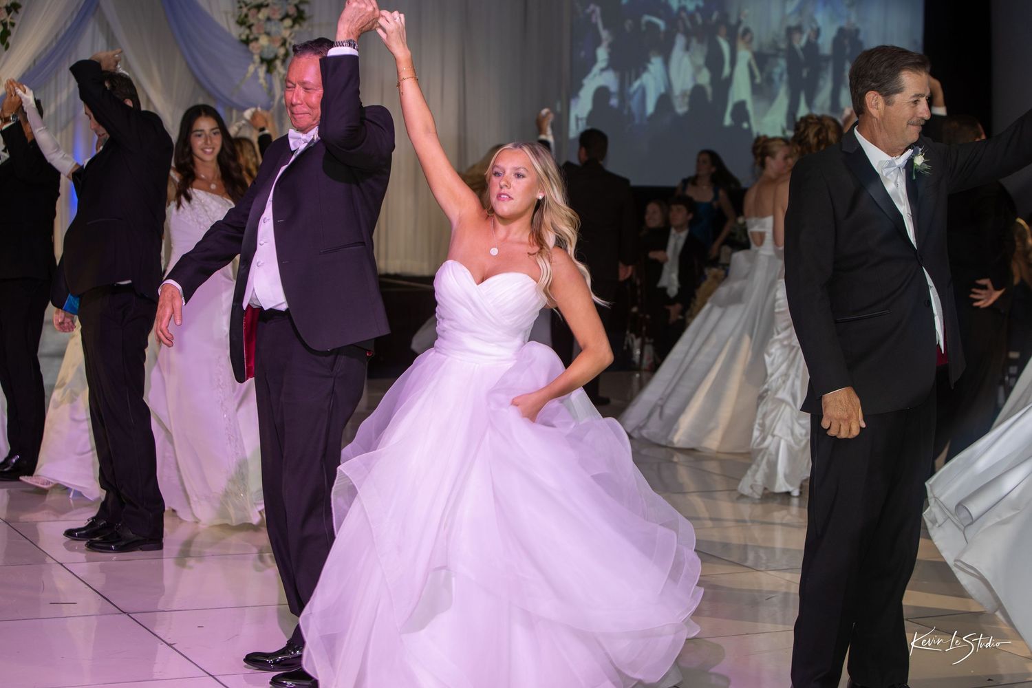 Wedding guests dance and celebrate on a dimly lit dance floor illuminated by pink and purple lighting.