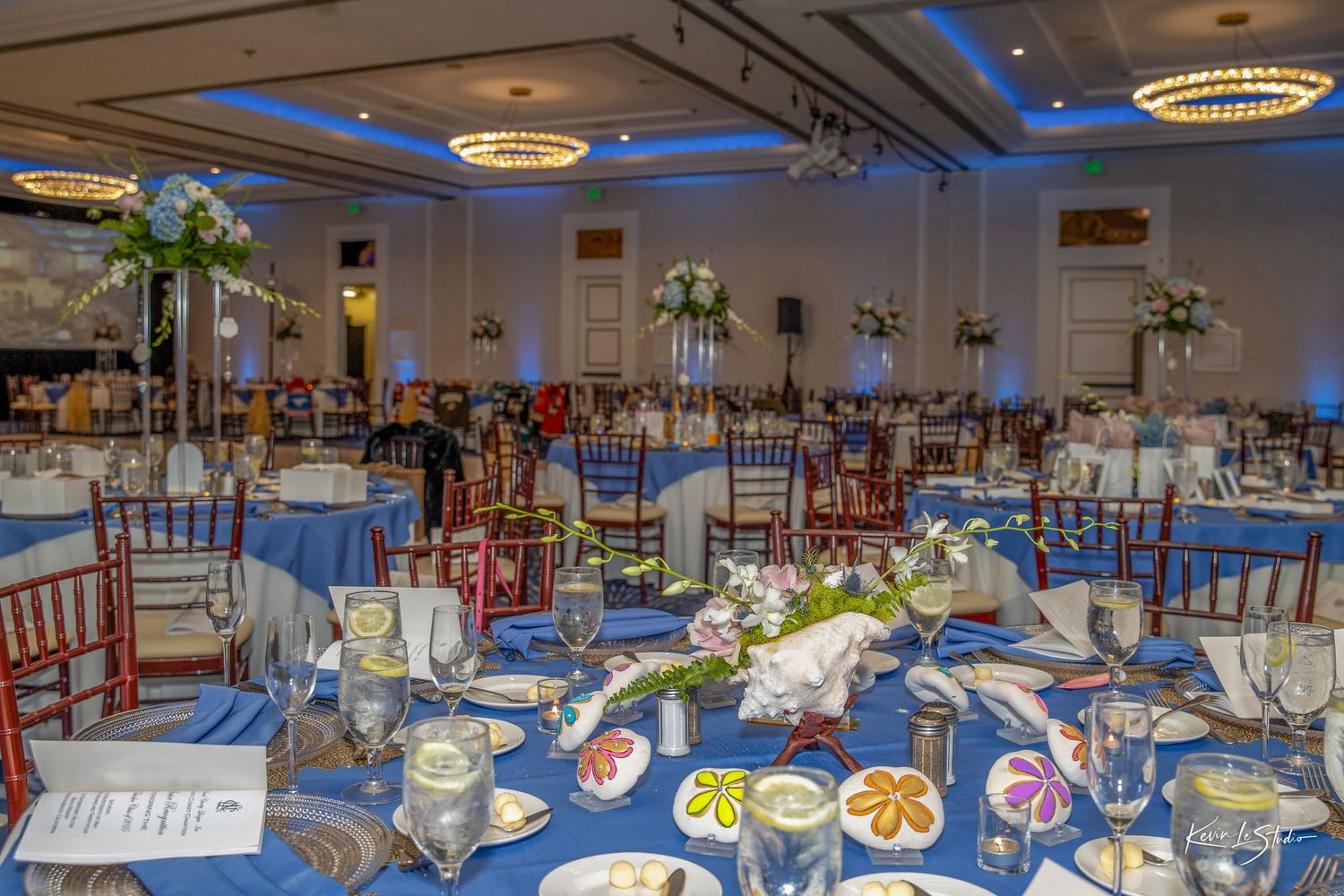 Elegant wedding reception hall with blue lighting, round tables, white linens, and ornate floral centerpieces.