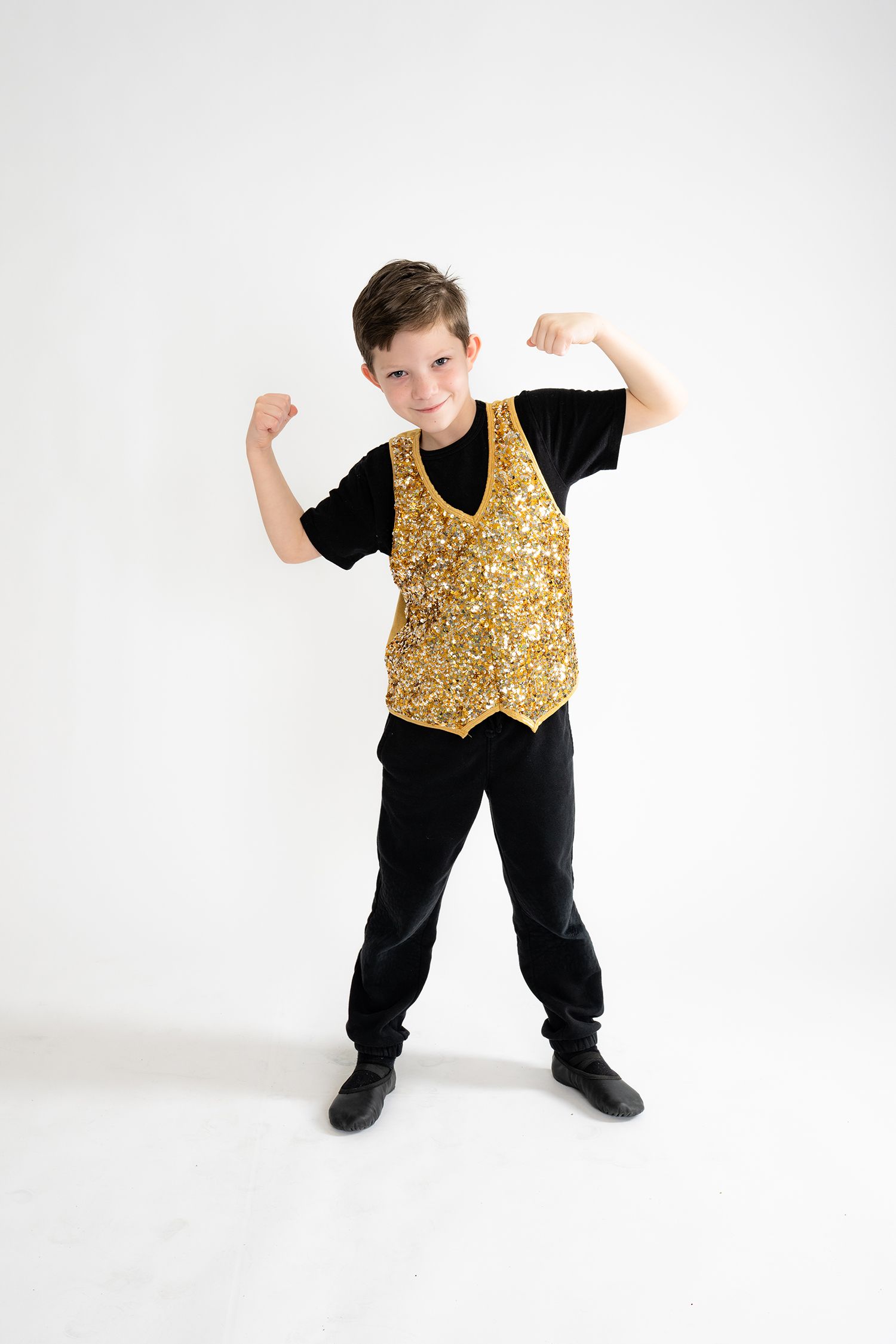 Young boy in gold sequin vest and black outfit flexing muscles, posing confidently on white background.
