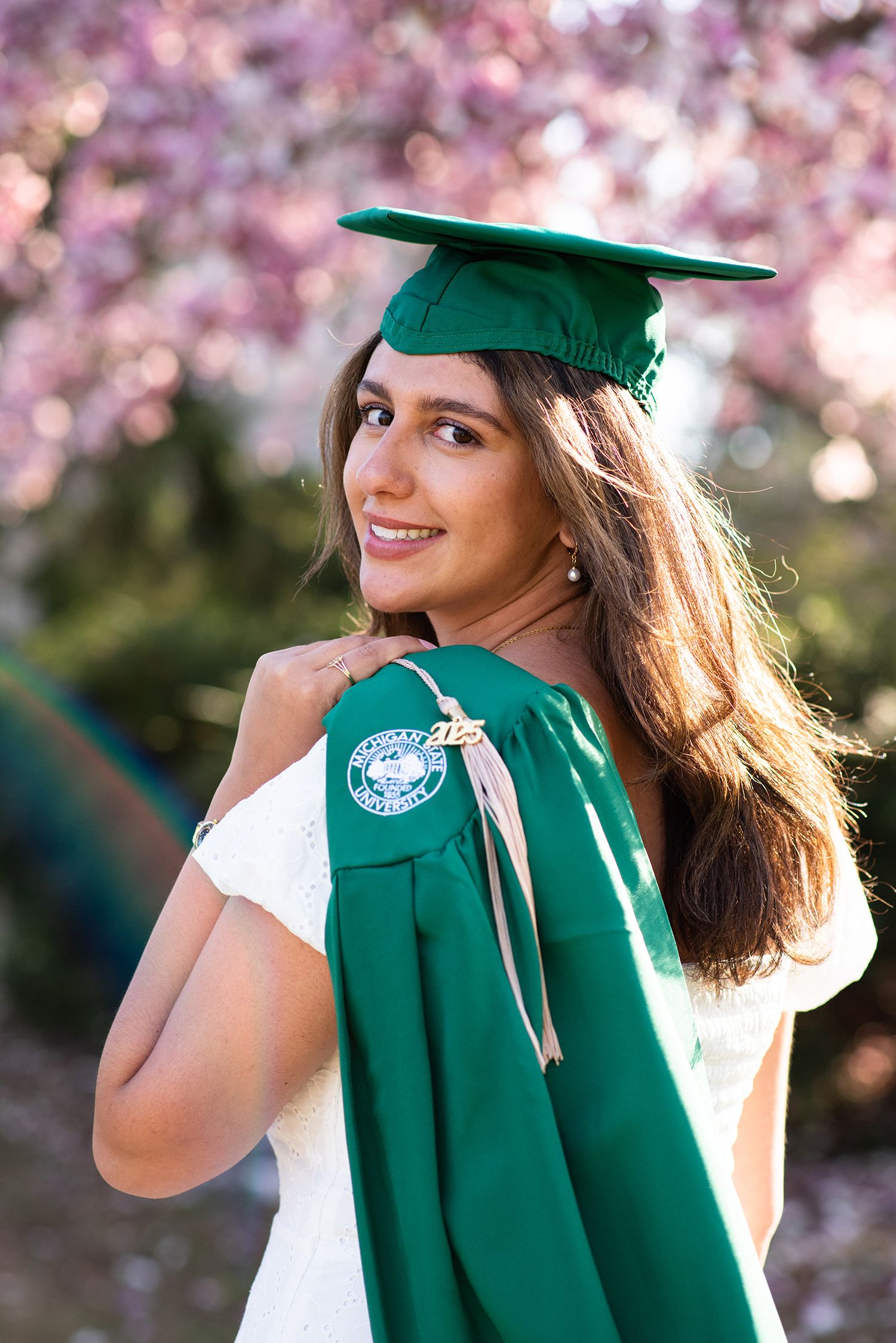Graduate in green cap and gown poses with cherry blossoms in spring.