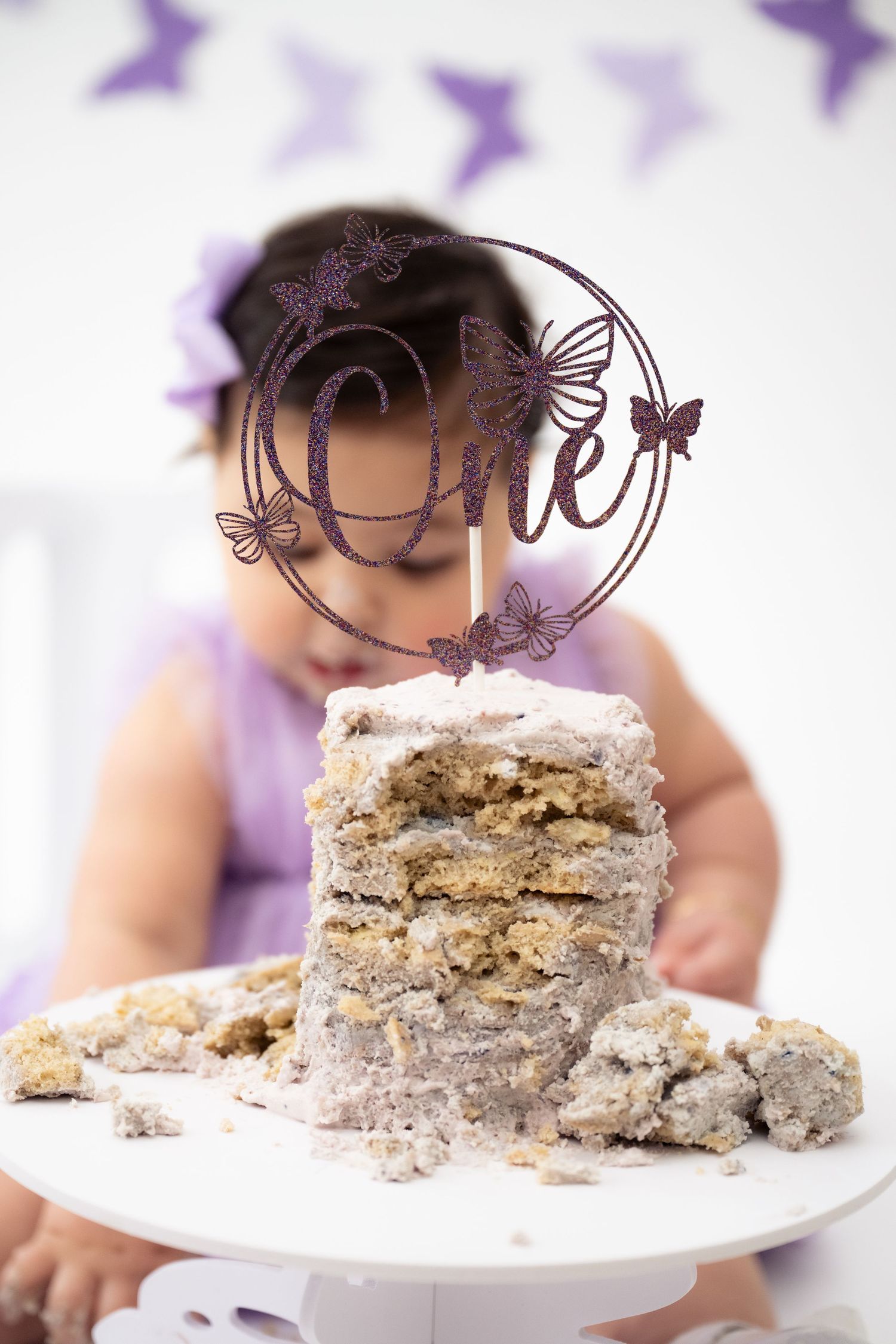 A white birthday cake with multiple layers sits on a cake stand with purple butterflies in the background.