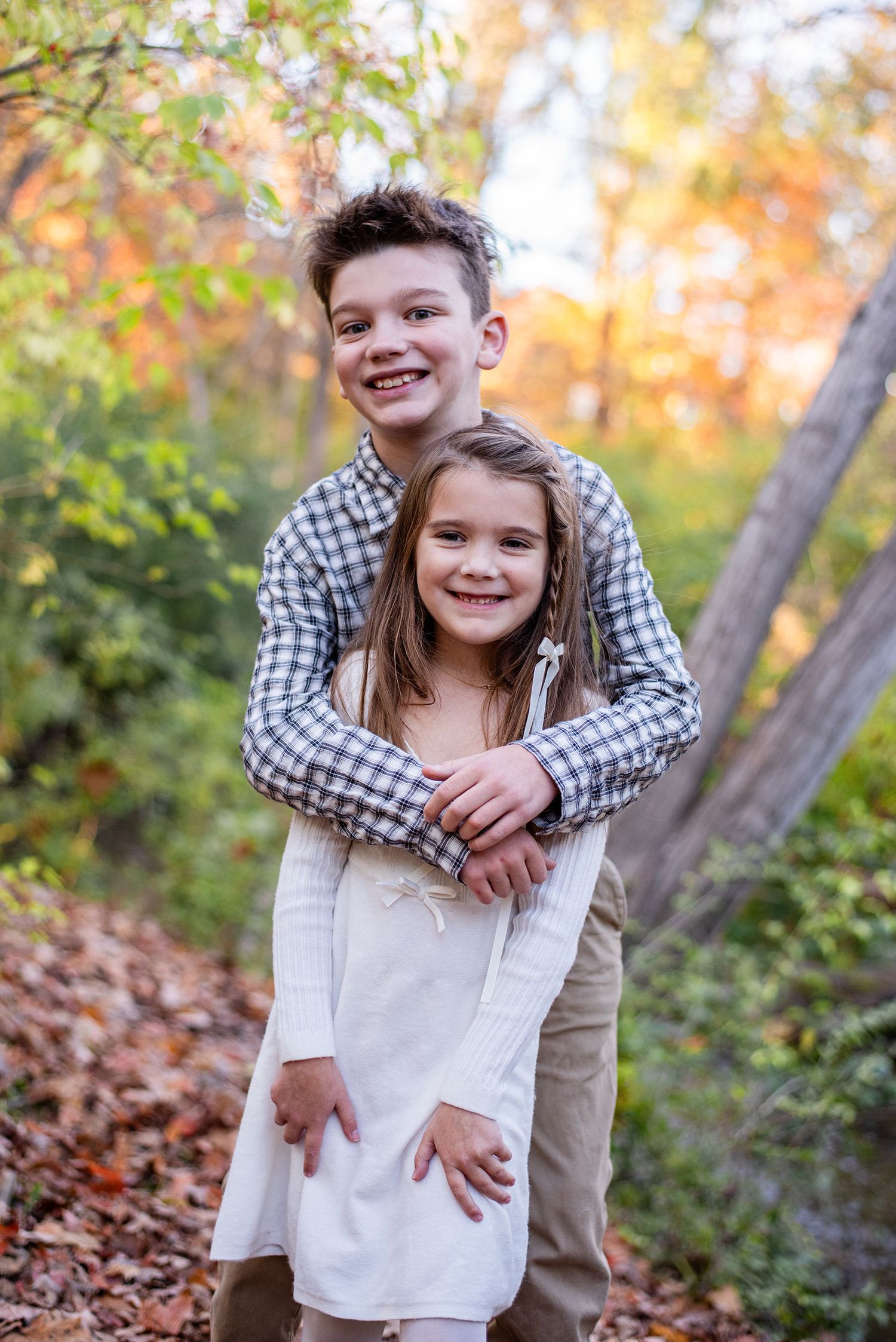 Two children playfully posing together in autumn leaves with warm golden light filtering through trees.