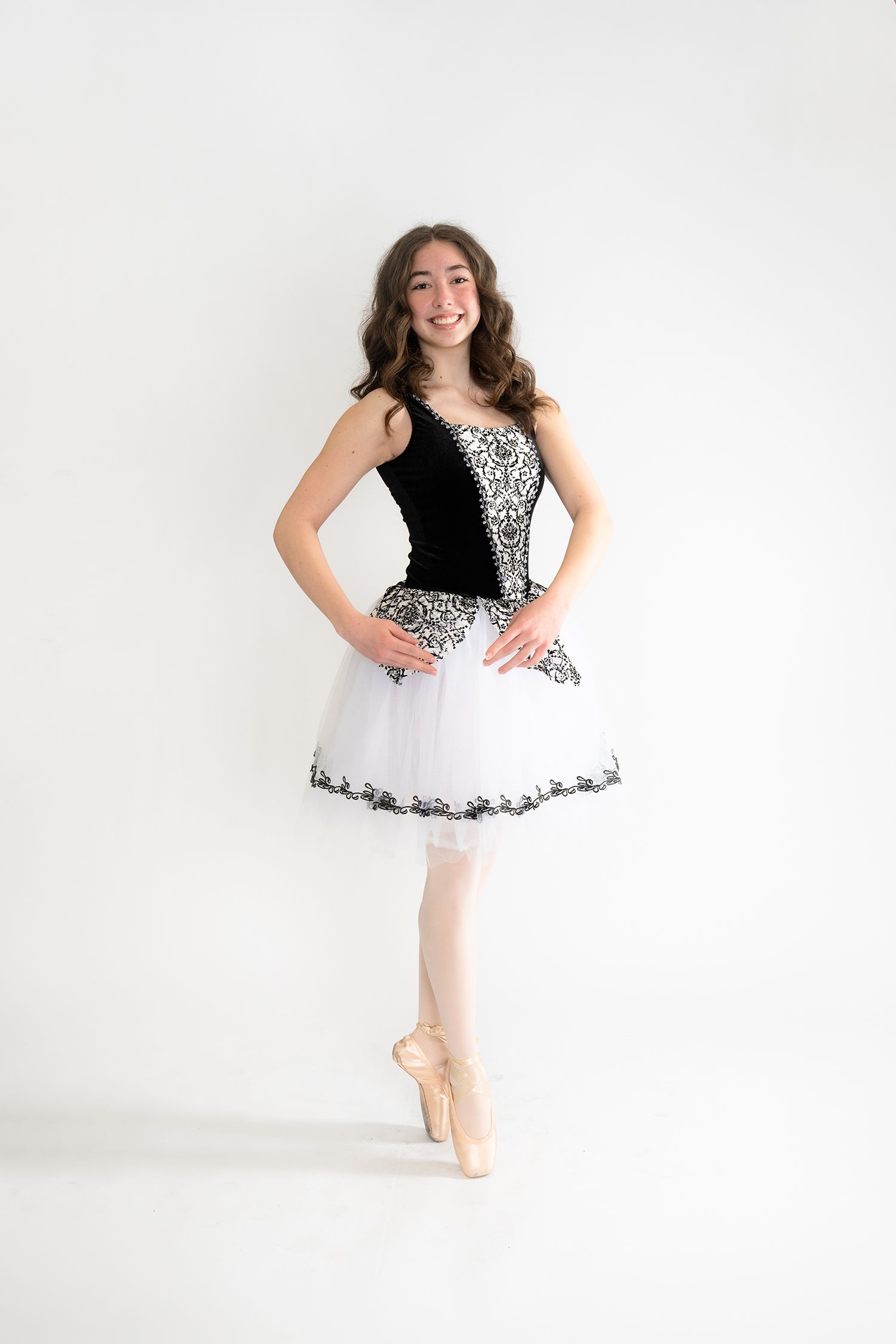 Young ballerina in black and white tutu costume poses en pointe, smiling against white background.