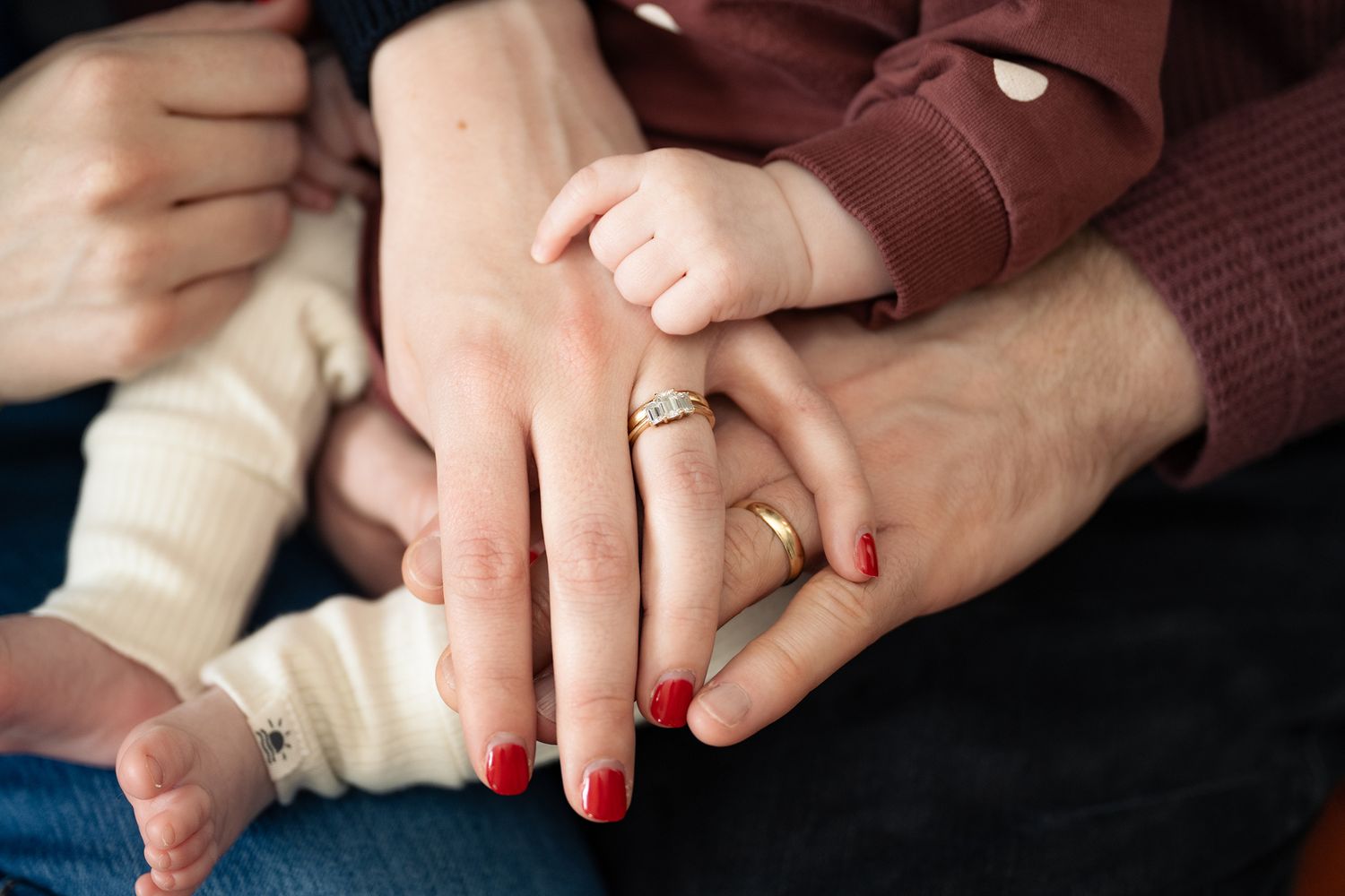Close-up of hands with red nail polish and engagement ring intertwined against dark background.