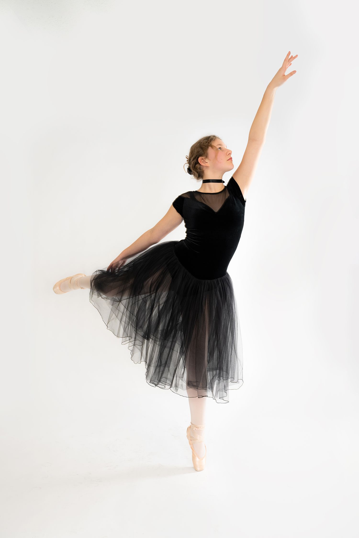 A young ballerina in a black tutu and pointe shoes performs an arabesque pose against a white background.
