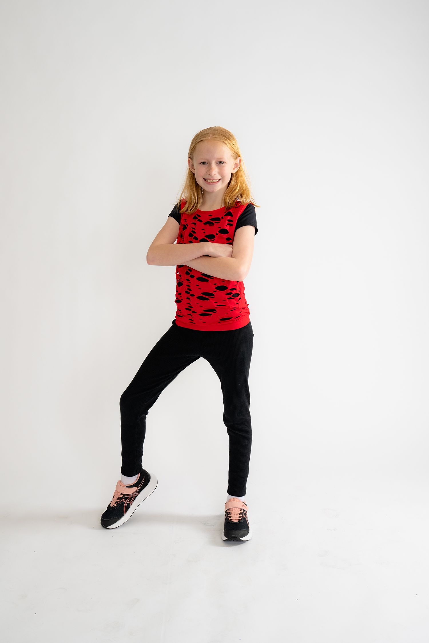 A young redhead girl poses confidently with crossed arms, wearing a red patterned top, black leggings, and sneakers.