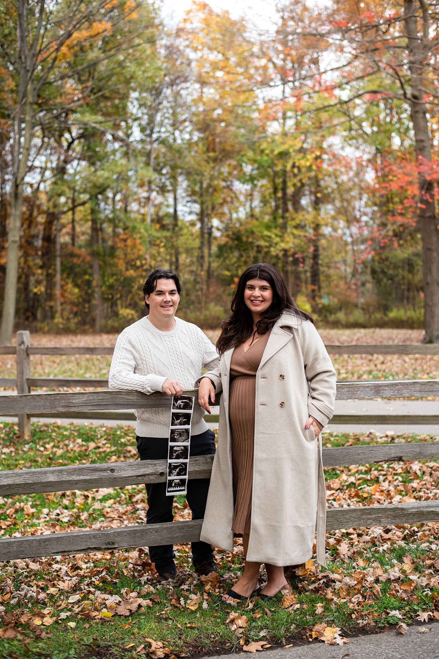 Couple posing with ultrasound photos in autumn park setting with fallen leaves.