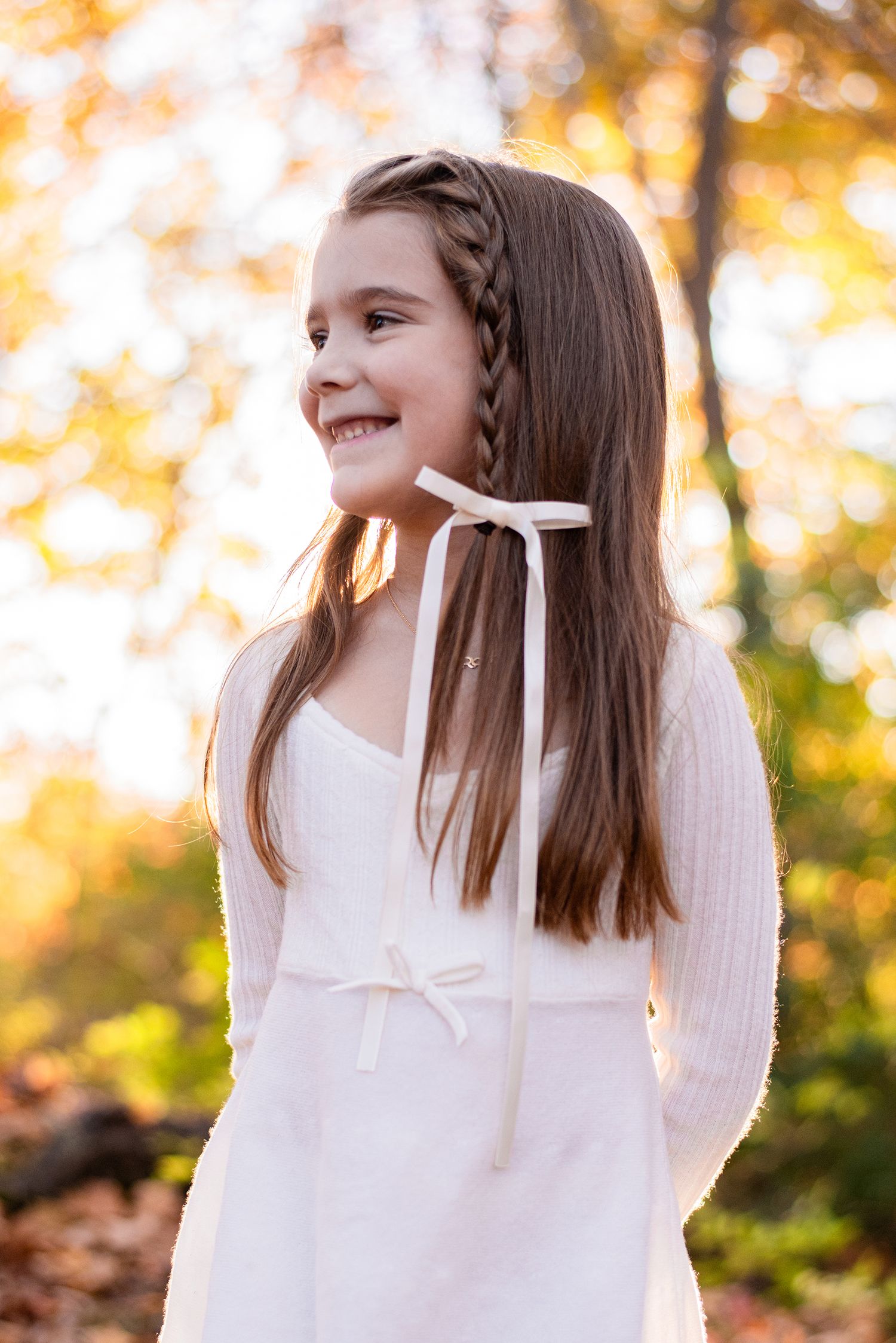 Girl in white dress posing against autumn foliage background with soft golden lighting.