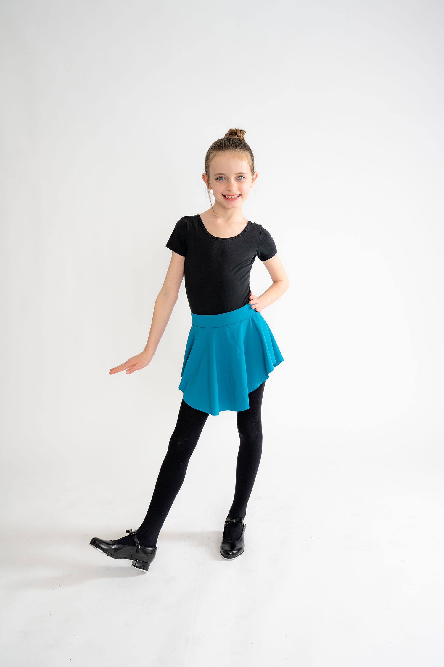 Young girl in black leotard, teal skirt, and tap shoes poses gracefully in a dance studio.