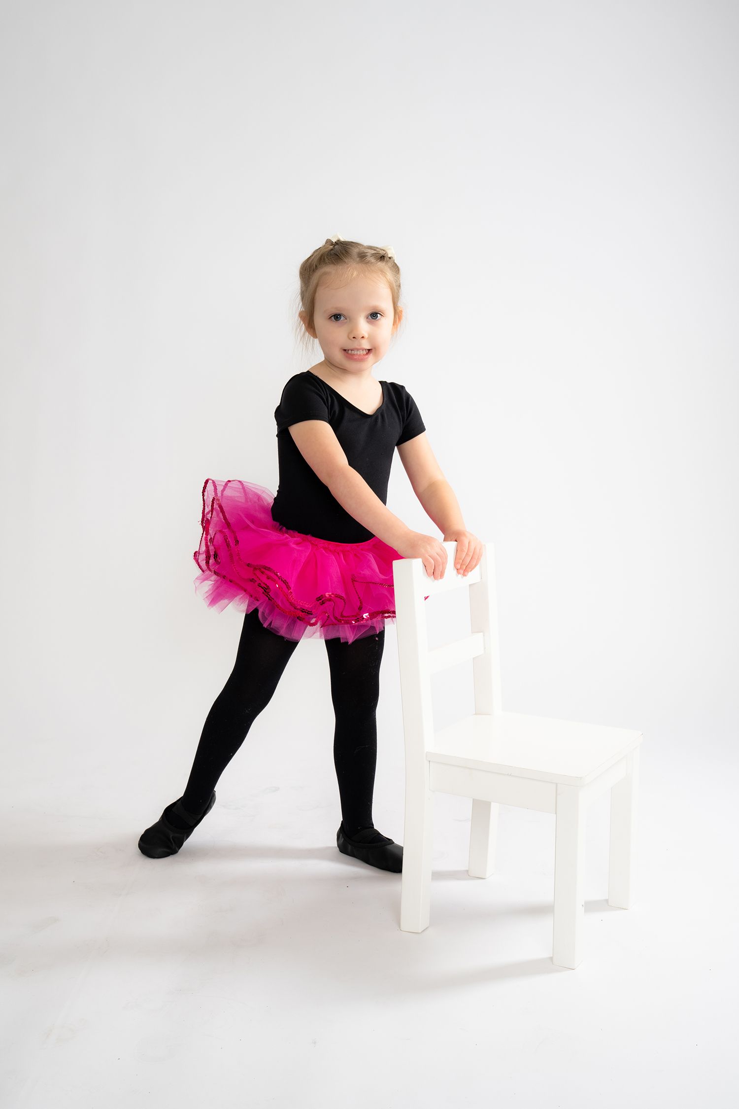 Young girl in black leotard and pink tutu standing beside a white chair on white background.