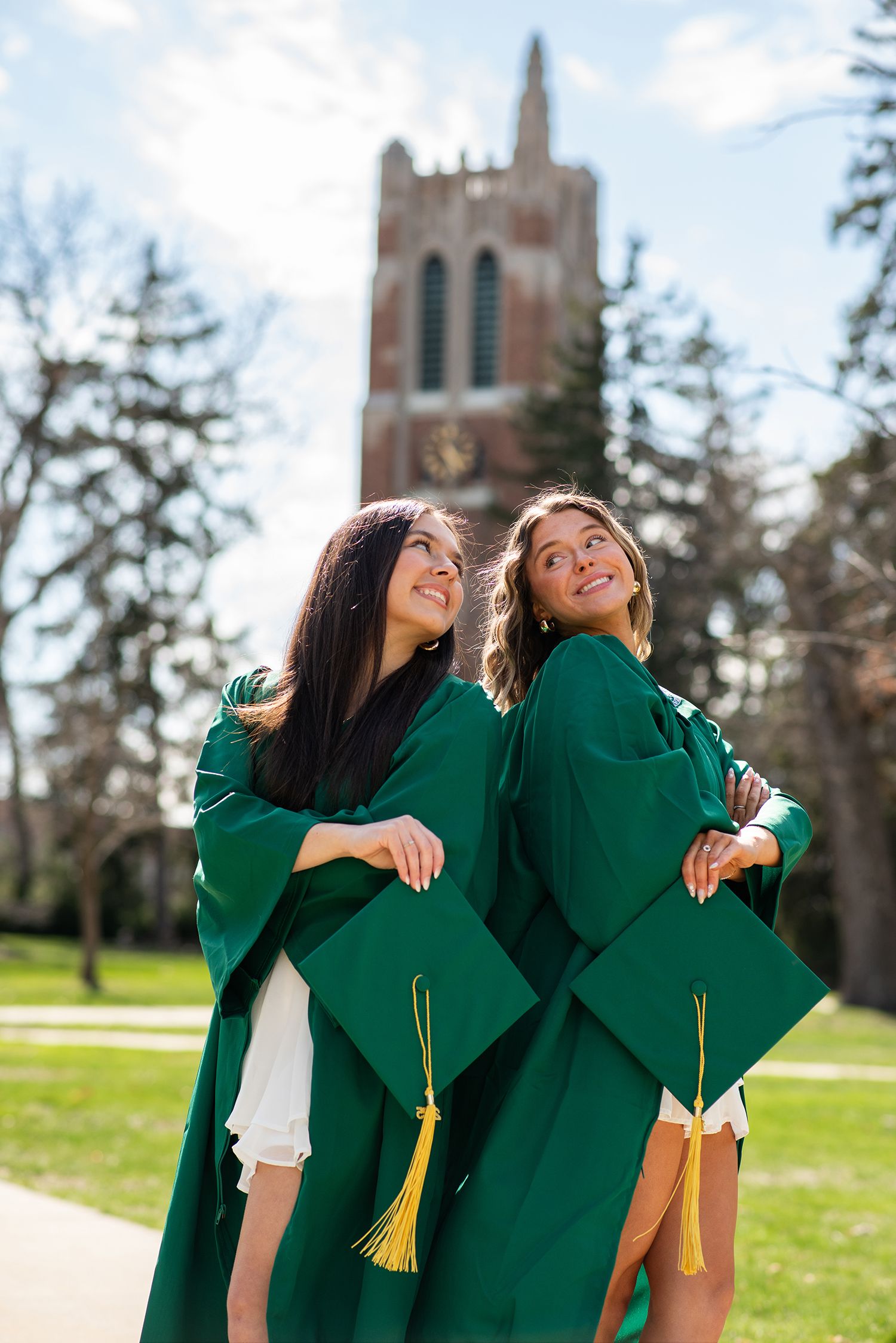 Graduates in green robes celebrate on campus lawn with gothic architecture in background.