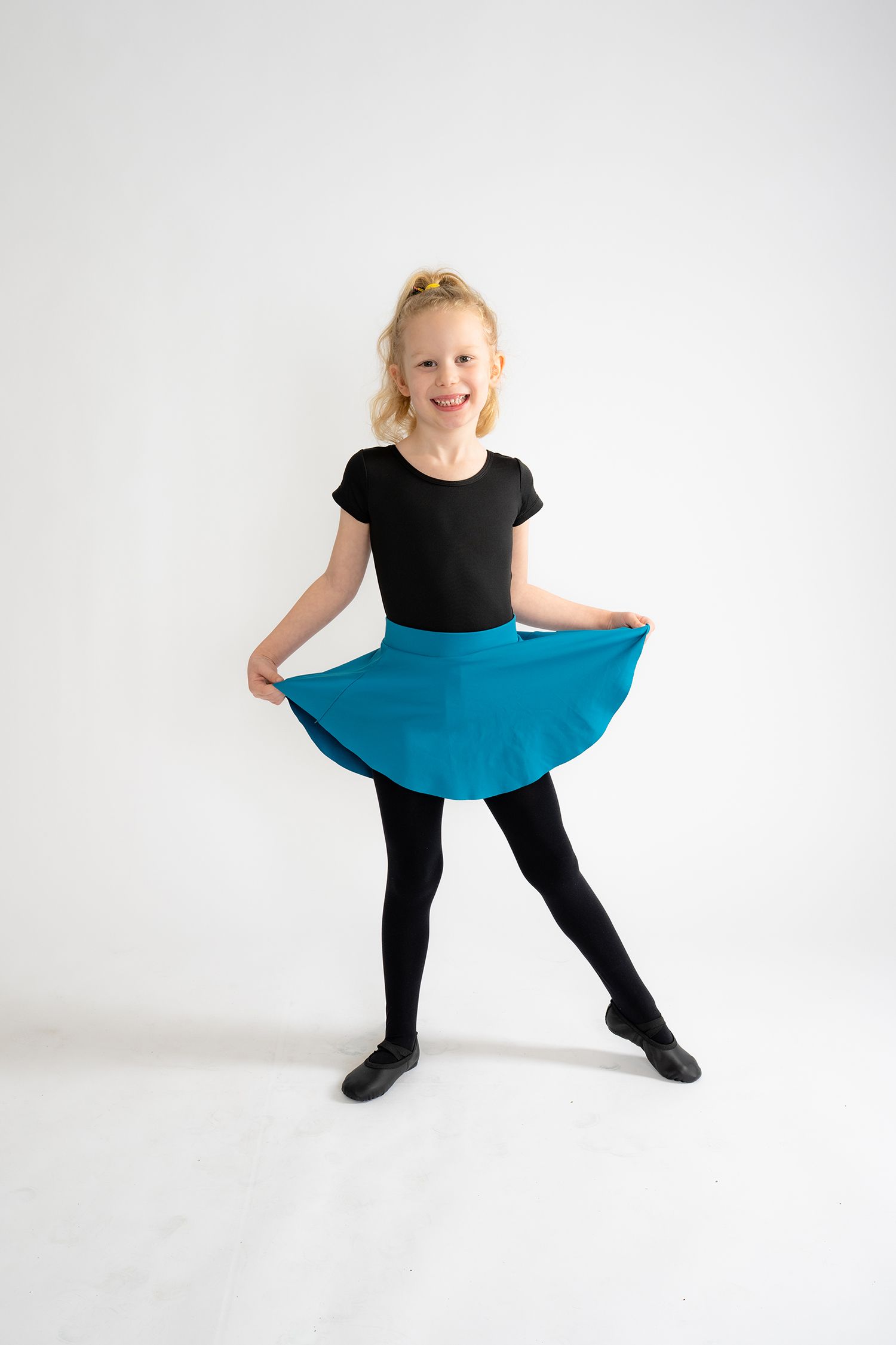 Young blonde girl in black leotard and teal skirt poses in ballet stance, smiling in dance studio.