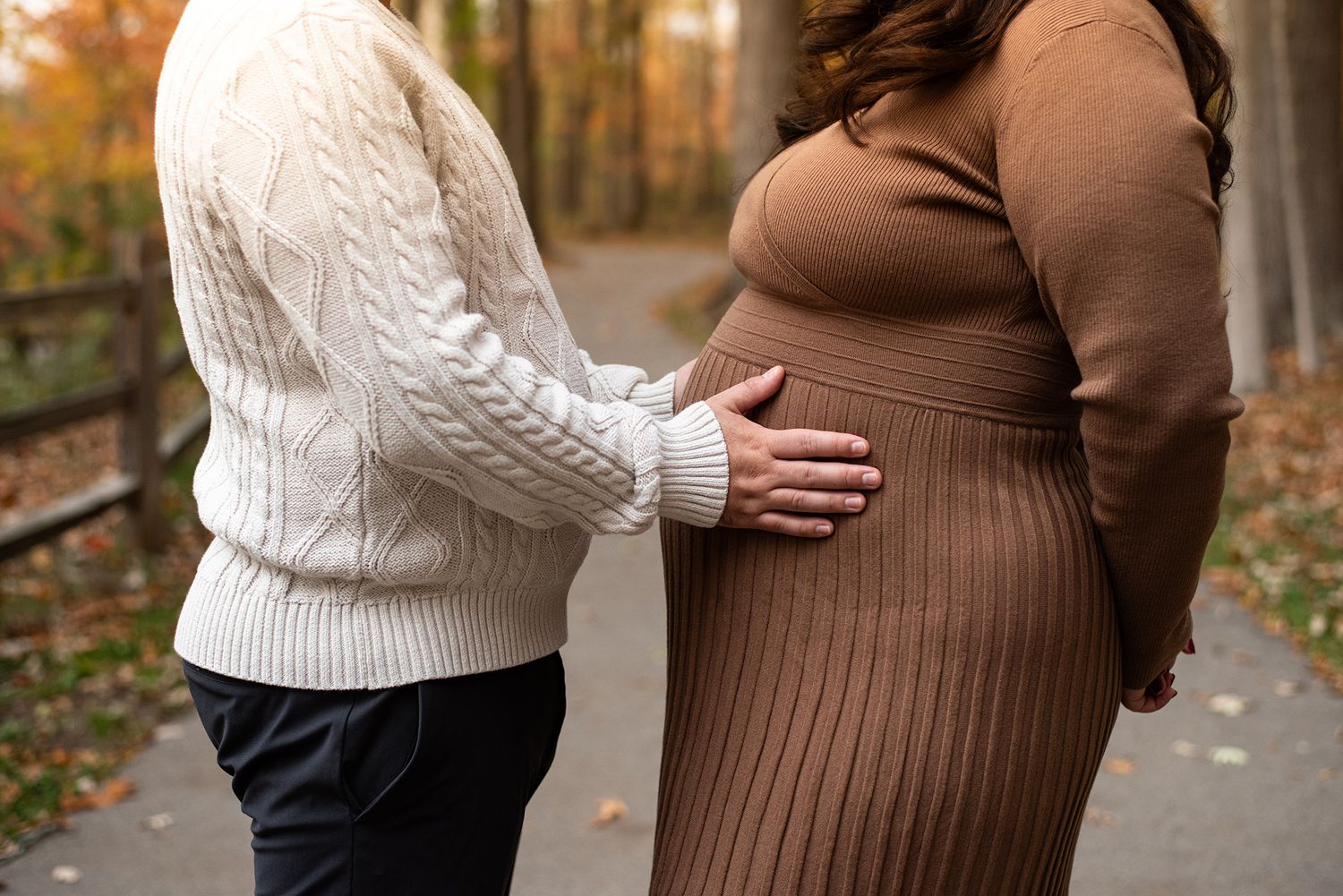 Maternity photo shoot showing couple in white and brown sweaters embracing outdoors in autumn.