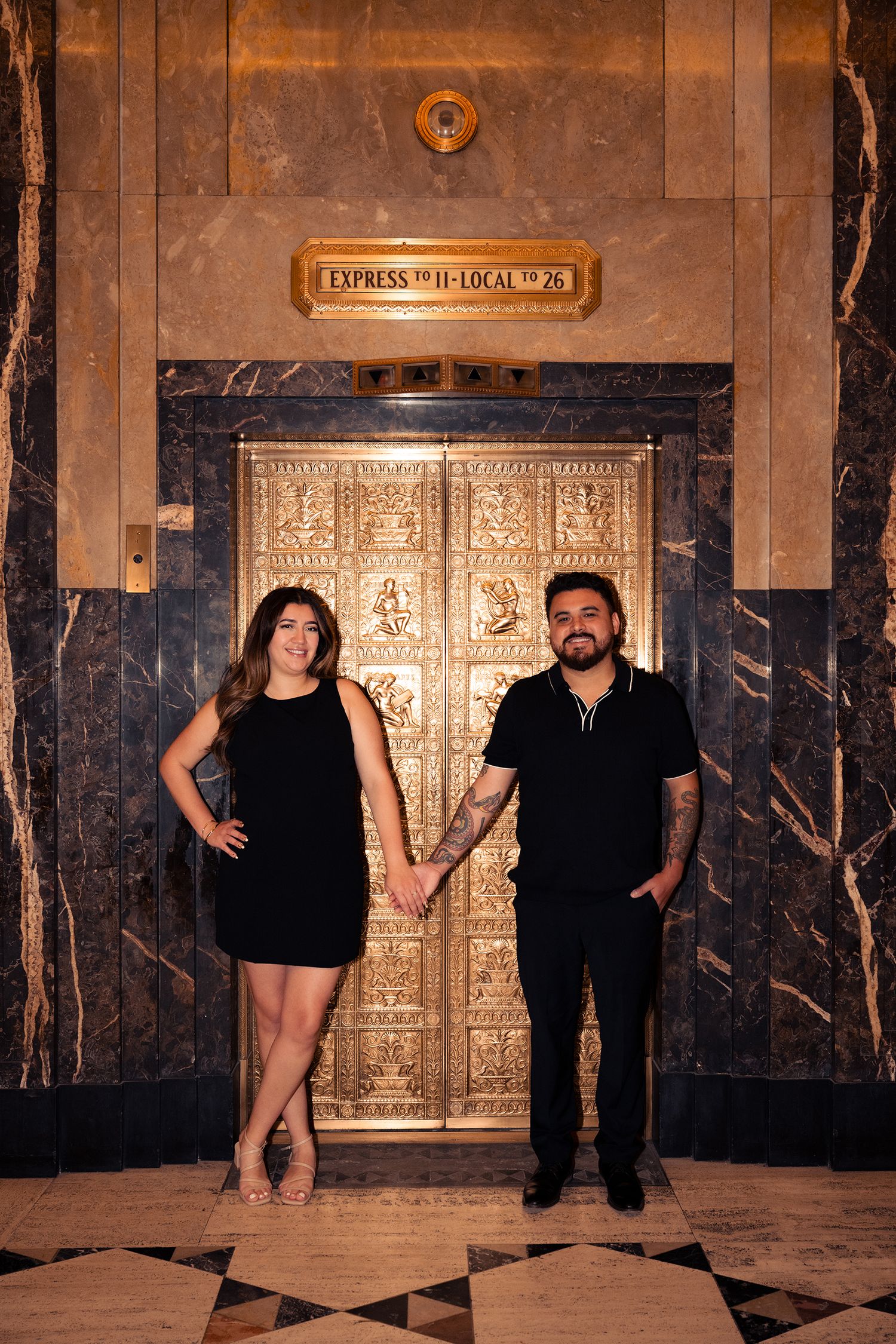 Couple in black outfits posing playfully against ornate art deco elevator doors.