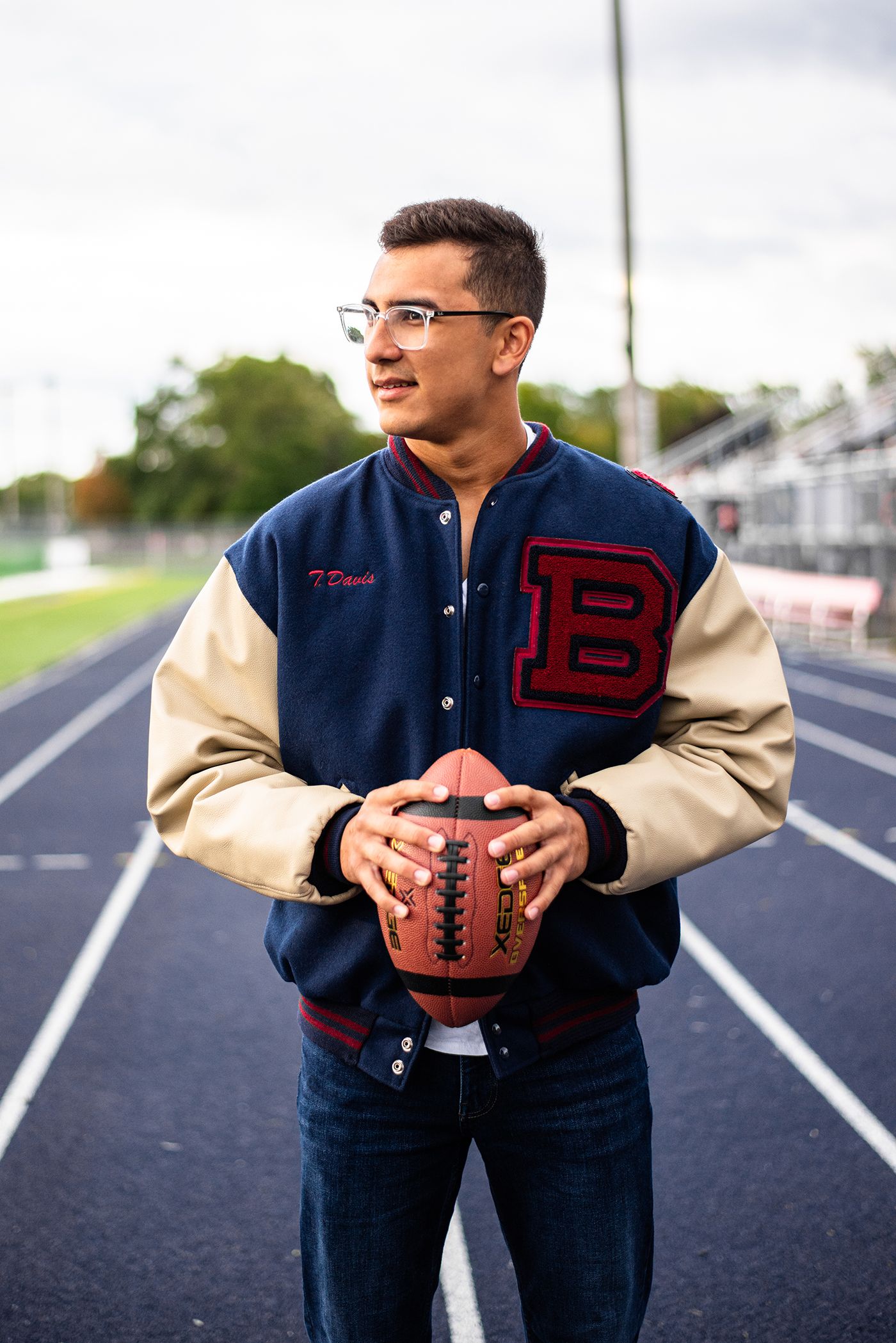 Student athlete in varsity jacket holding football on track field.