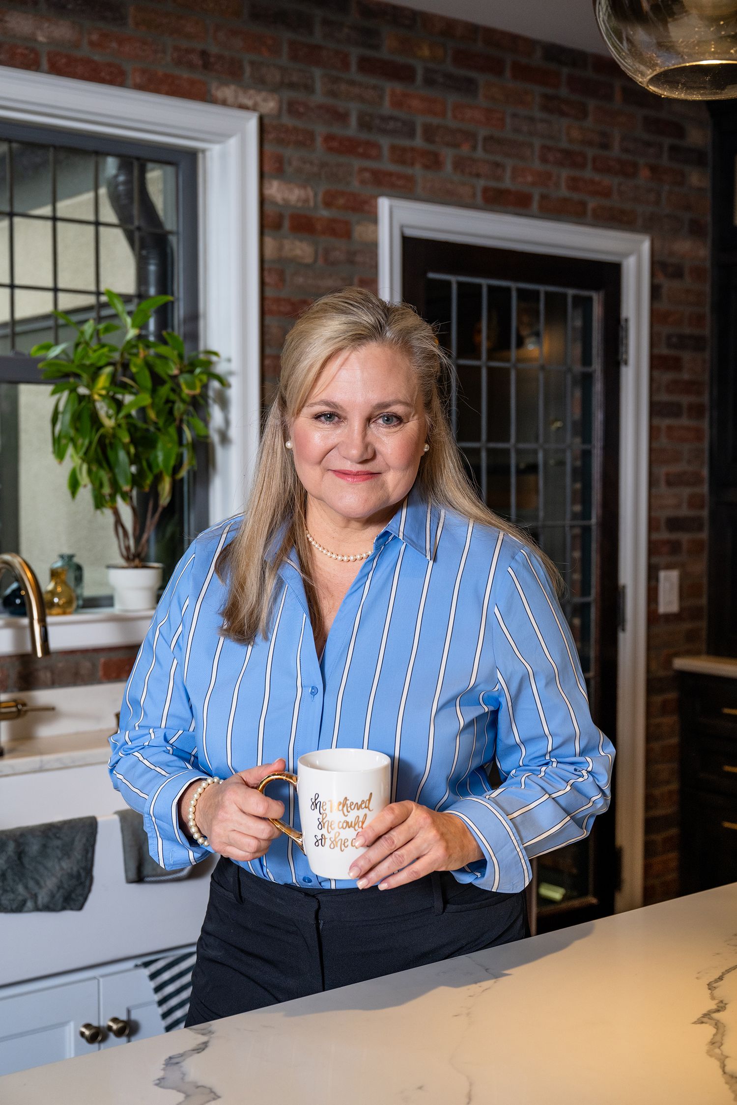 Professional woman in striped blue shirt sitting at counter with coffee mug in modern brick office.