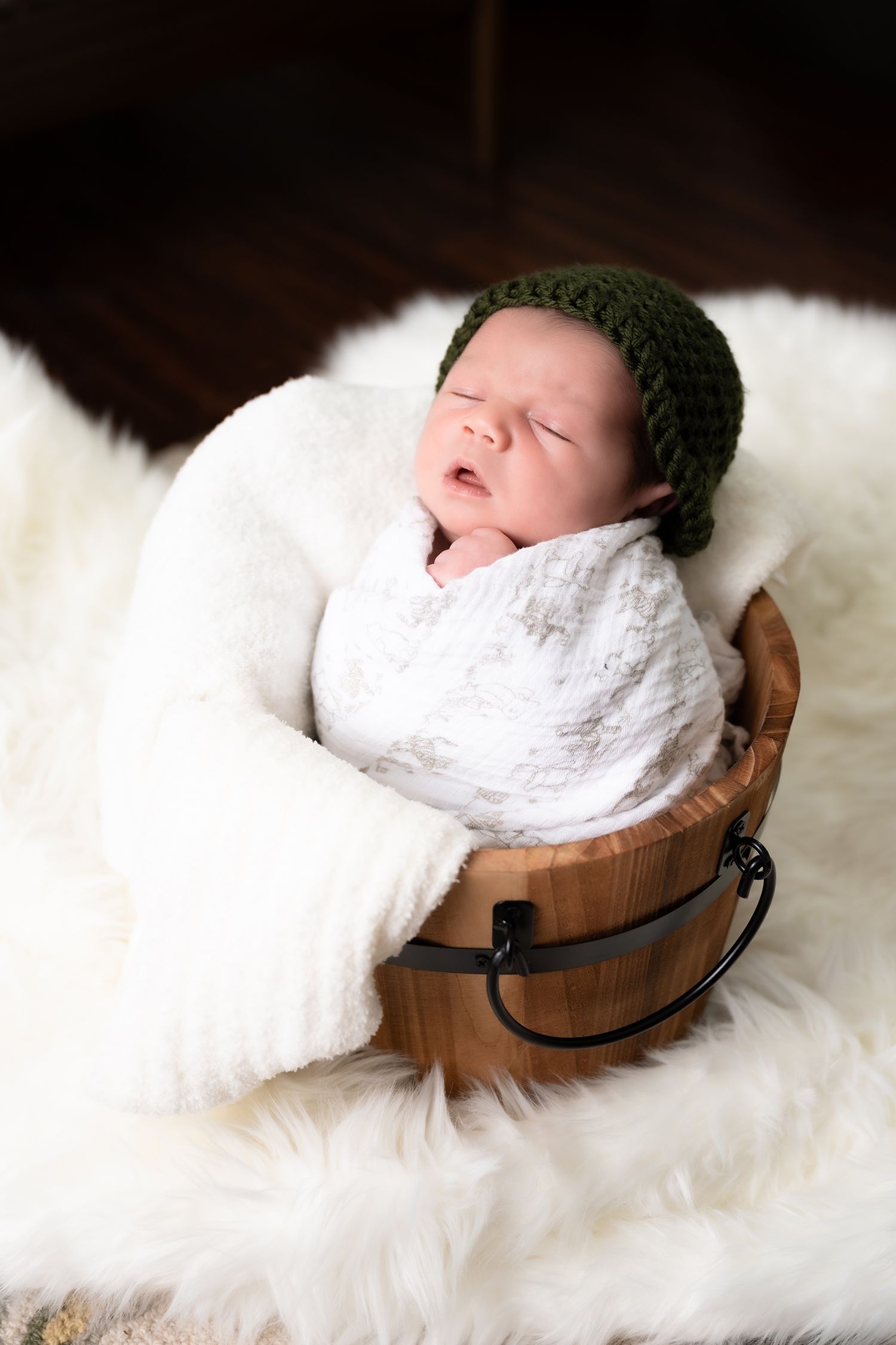 Sleeping newborn wrapped in white blanket lying in rustic wooden bowl on fluffy white fur backdrop.