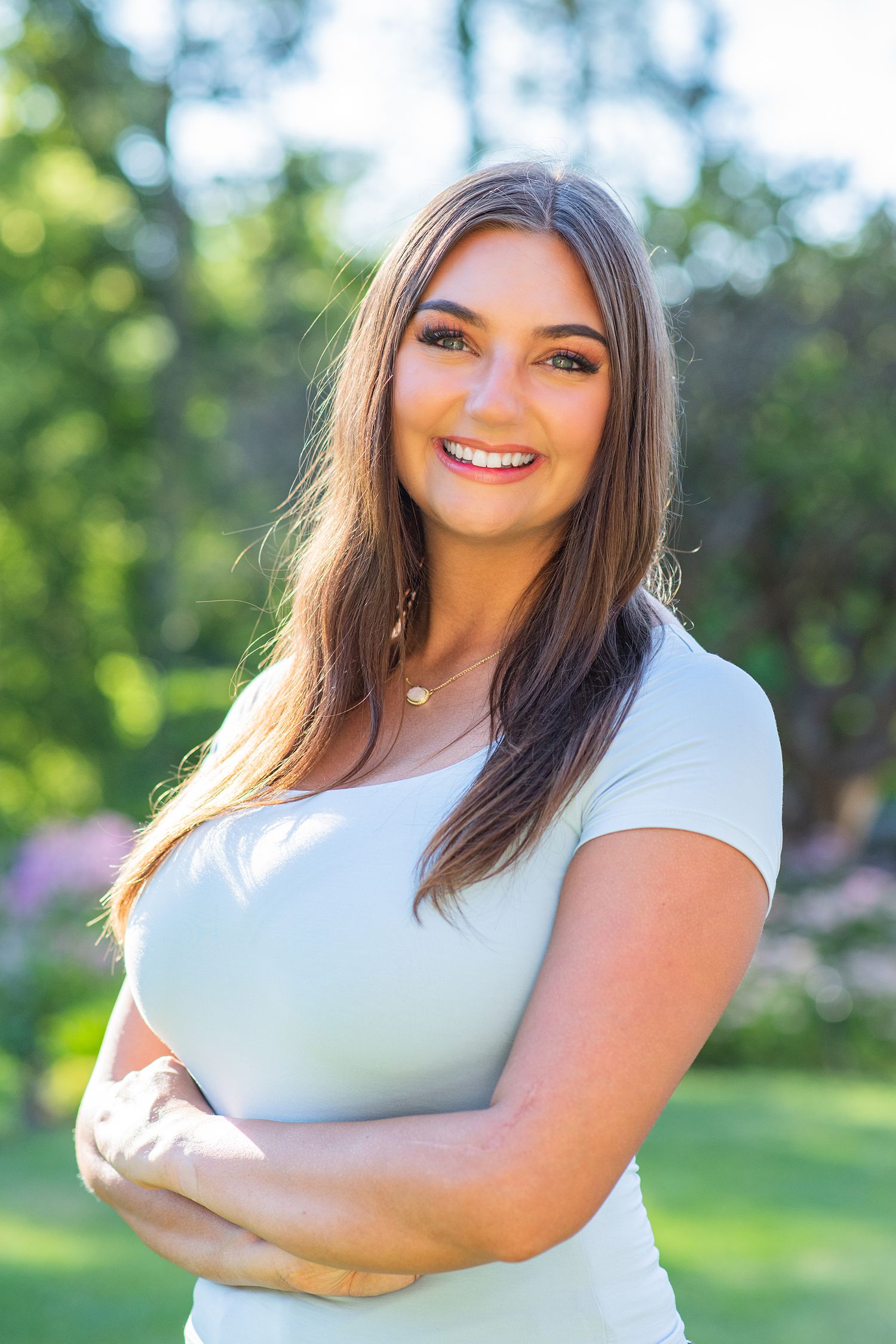 Young woman in white t-shirt posing outdoors with crossed arms and natural summer background.