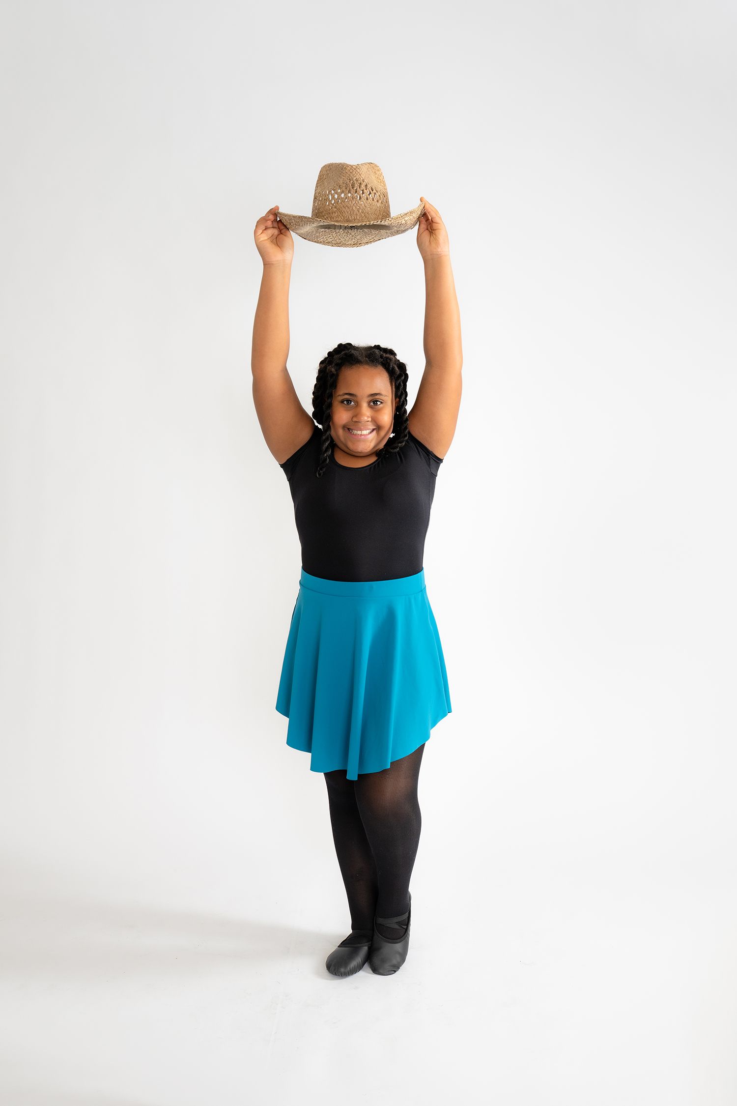 Young woman in black top, teal skirt, and tights holds a straw cowboy hat above her head, smiling.