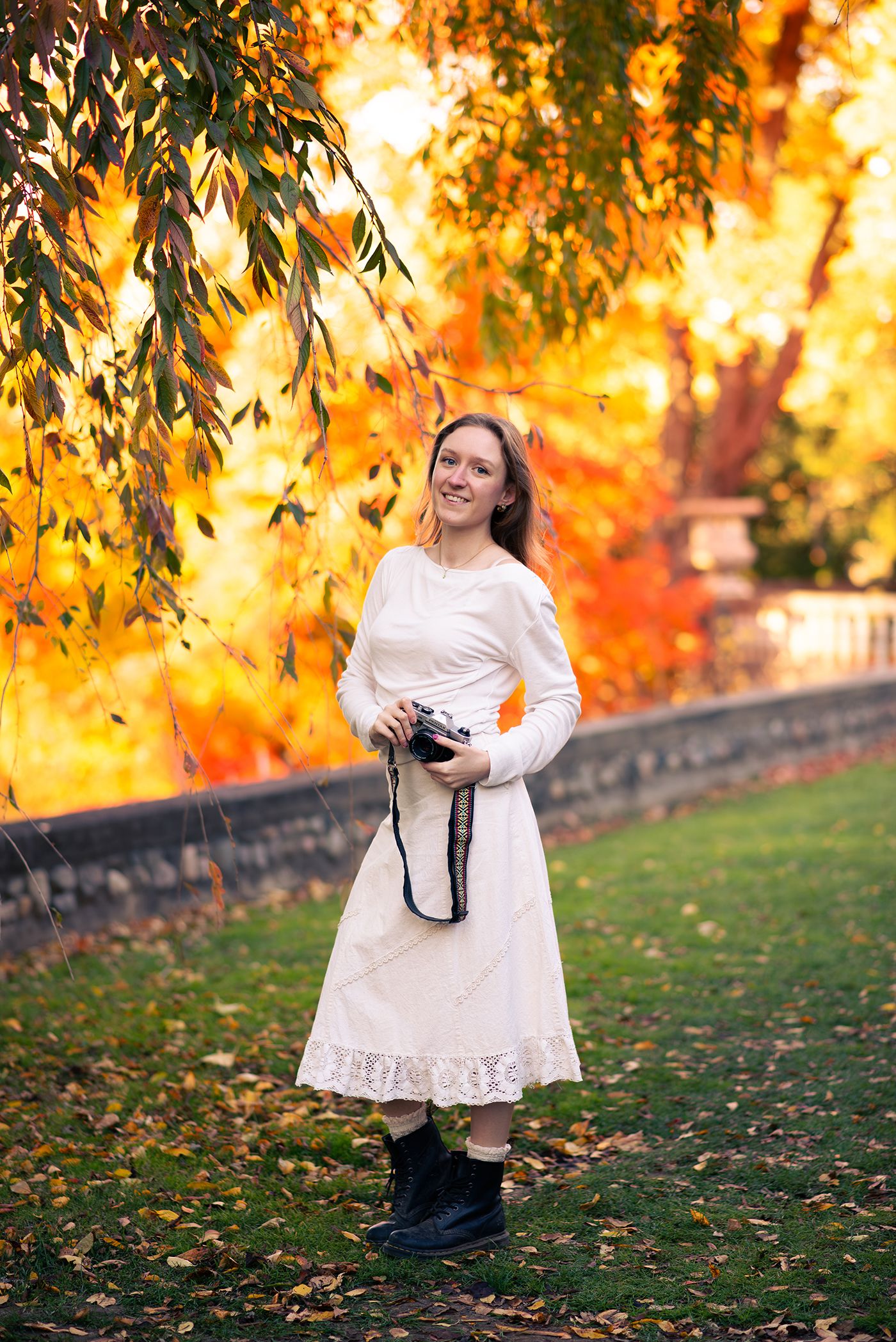 Person in white dress poses with camera against vibrant autumn foliage at sunset.
