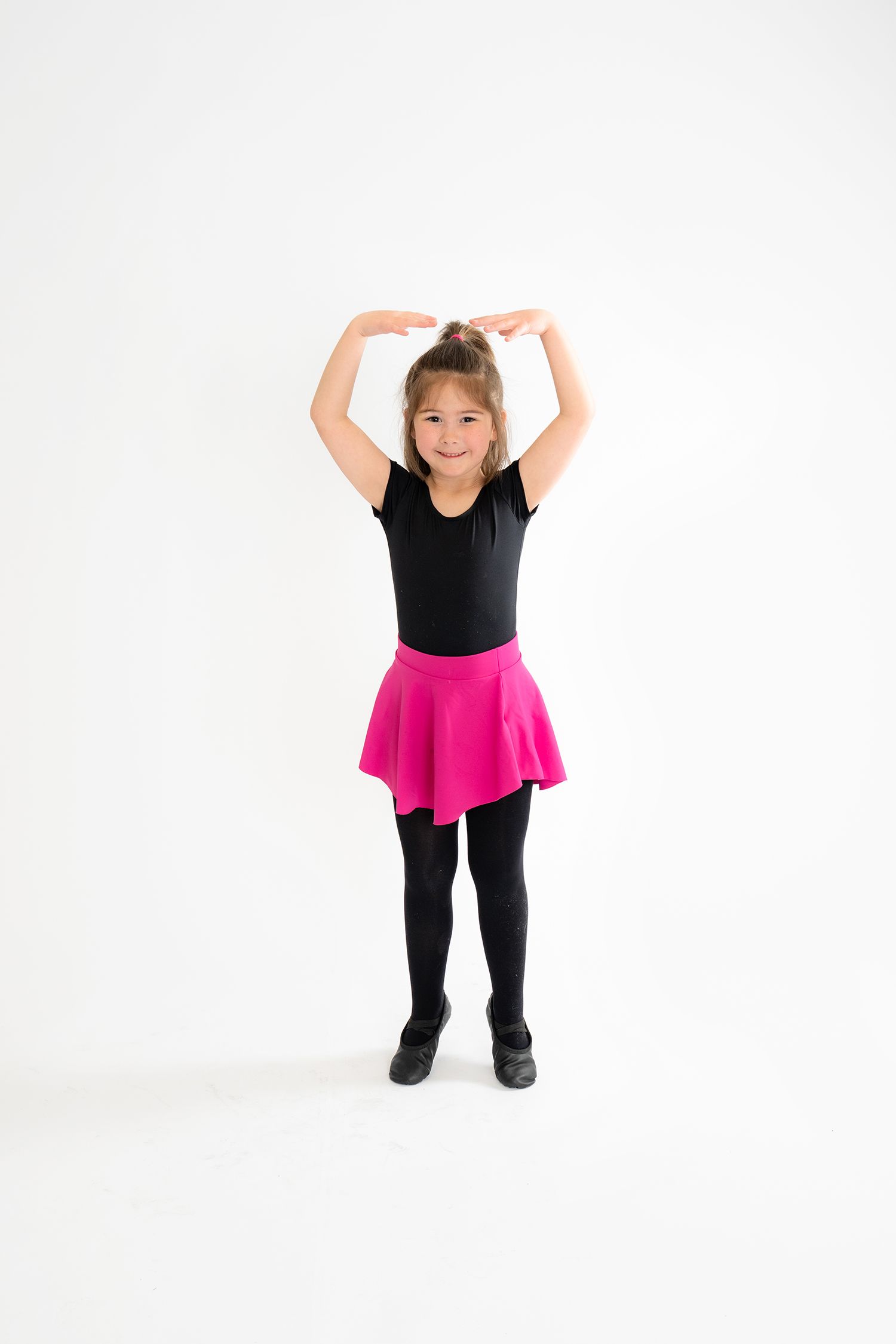 Young girl in black leotard and pink skirt poses with arms raised overhead in ballet dance position.