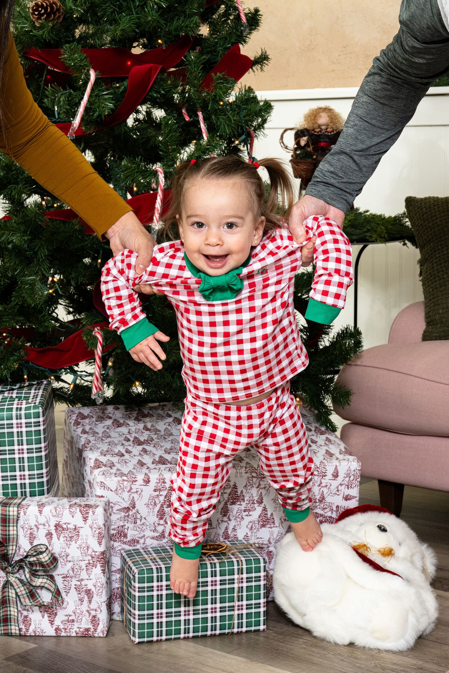 Toddler girl in red plaid Christmas pajamas standing by decorated tree with gifts, supported by parents hands.