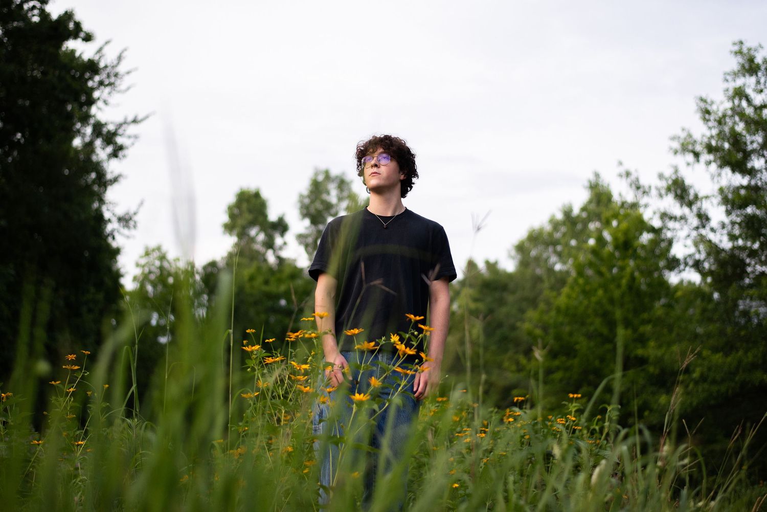 Person stands among tall wildflowers in summer field with cloudy sky.