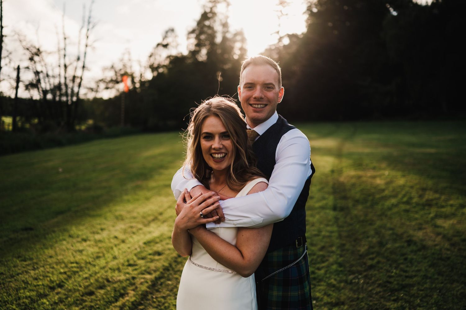 Romantic couple embraces in golden sunlight on grassy hillside during sunset photography session.