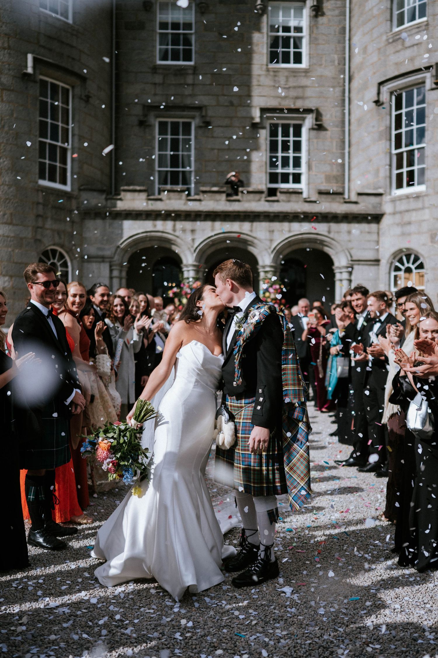 Wedding couple celebrating under confetti shower outside stone building with Scottish attire.