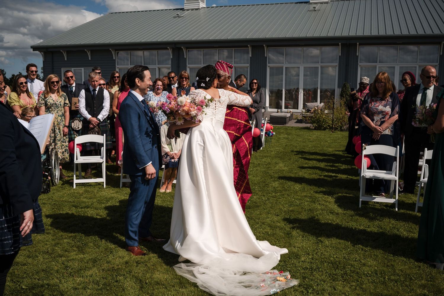 Guests seated in white chairs watch outdoor wedding ceremony on sunny day.