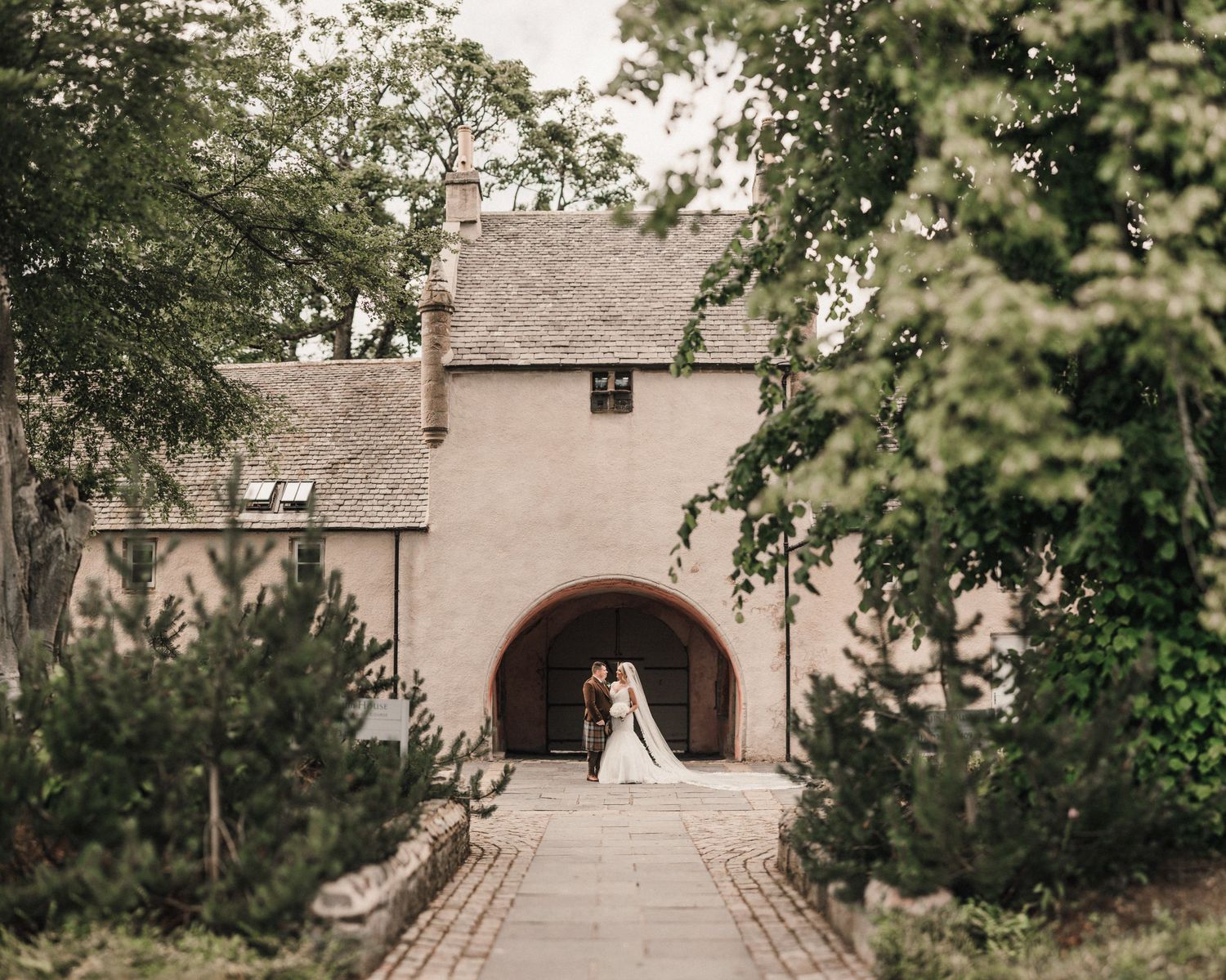 Stone archway entrance leads through tall trees and landscaped path.