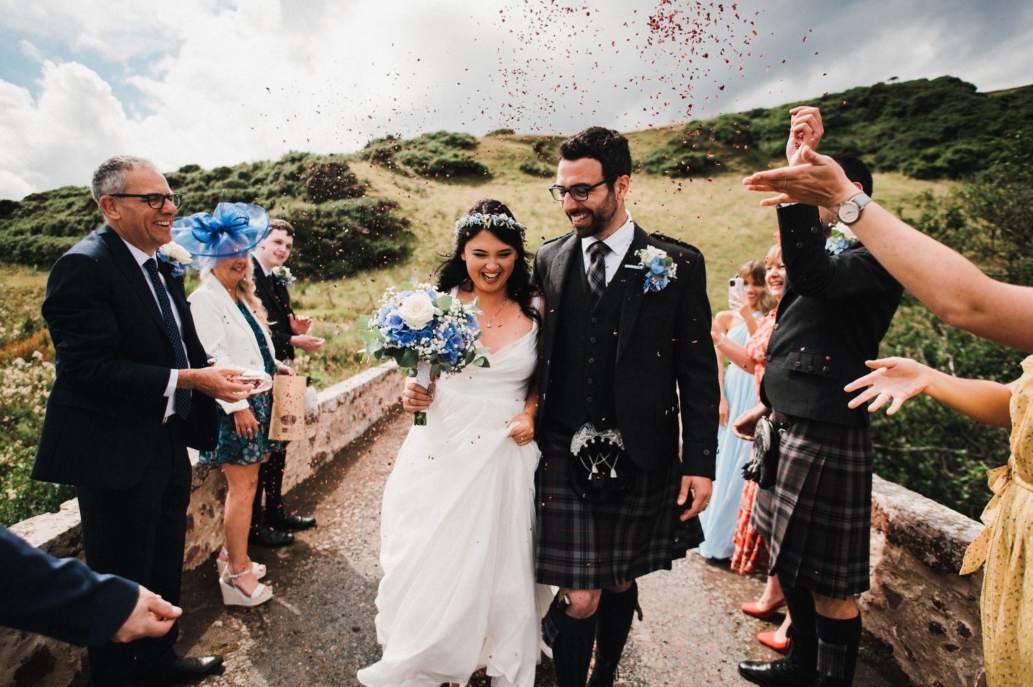 Wedding guests throw confetti as the newlyweds celebrate on a scenic Scottish hillside path.
