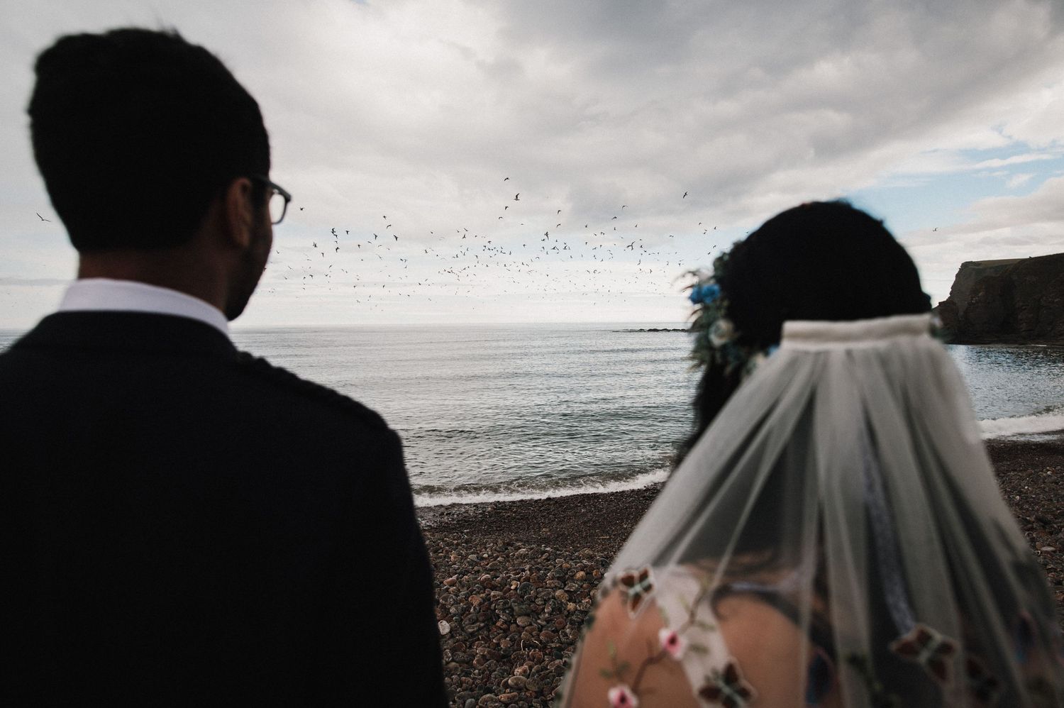 A silhouetted wedding couple gazes at seabirds flying over a coastal landscape.