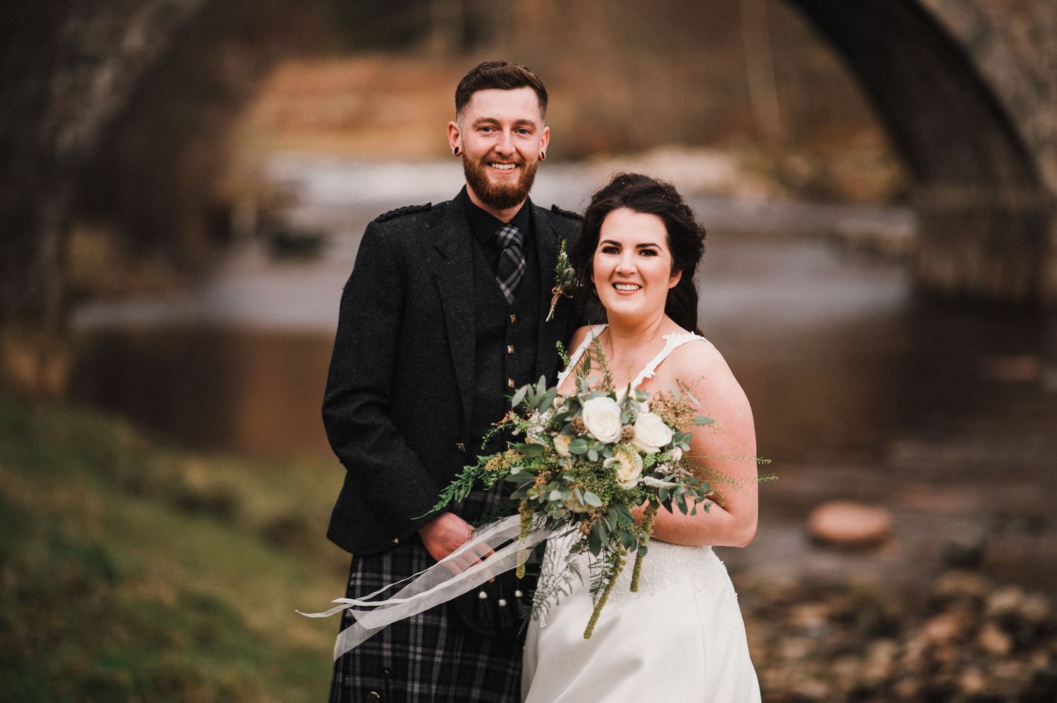Scottish wedding couple poses by stone bridge with bride holding rustic bouquet.