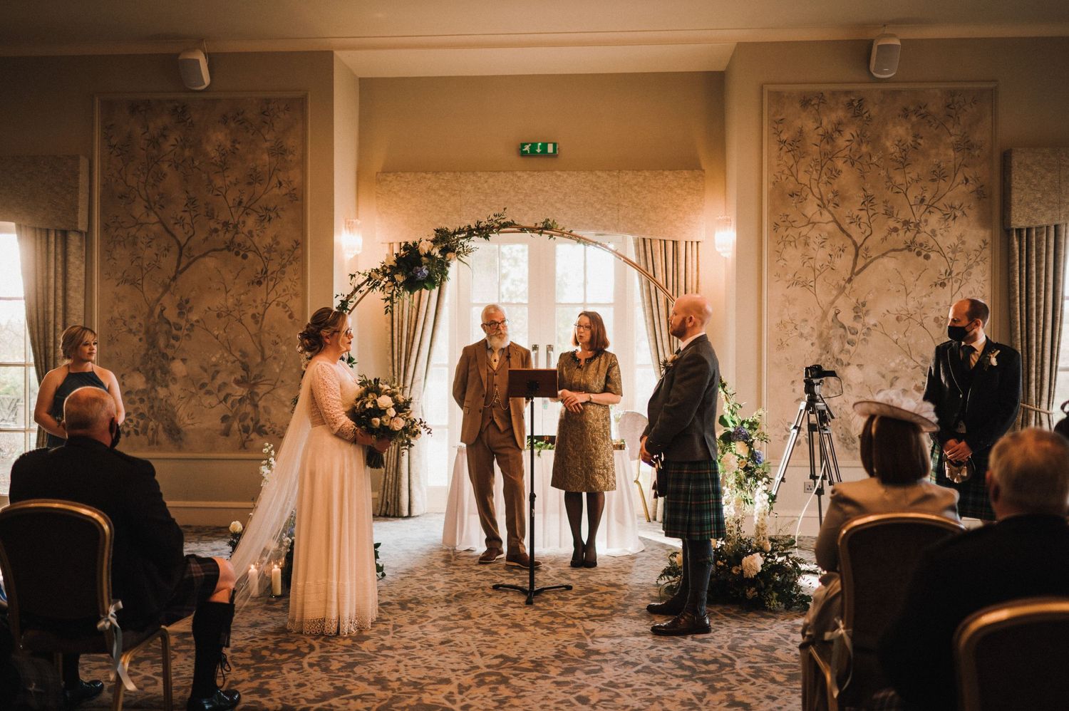 Wedding ceremony in an elegant room with patterned wallpaper and floral arch decorations.