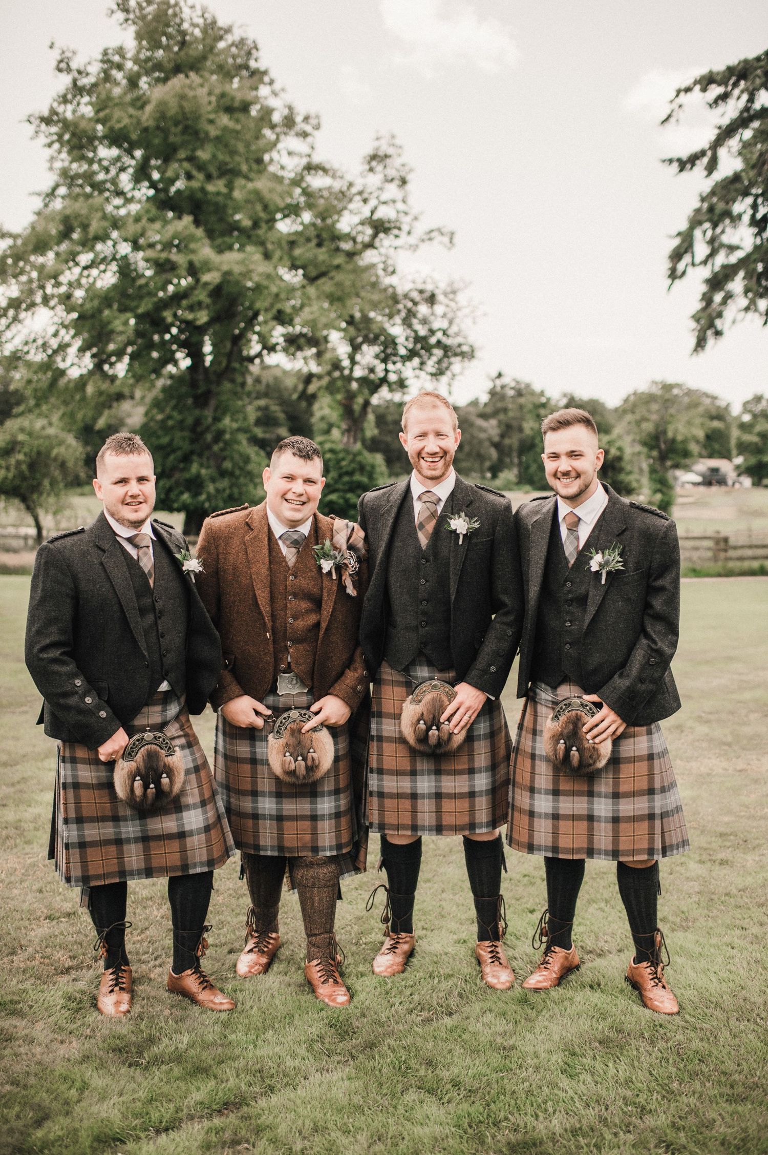 Four groomsmen in traditional Scottish kilts and jackets pose together on grassy lawn for wedding photo.