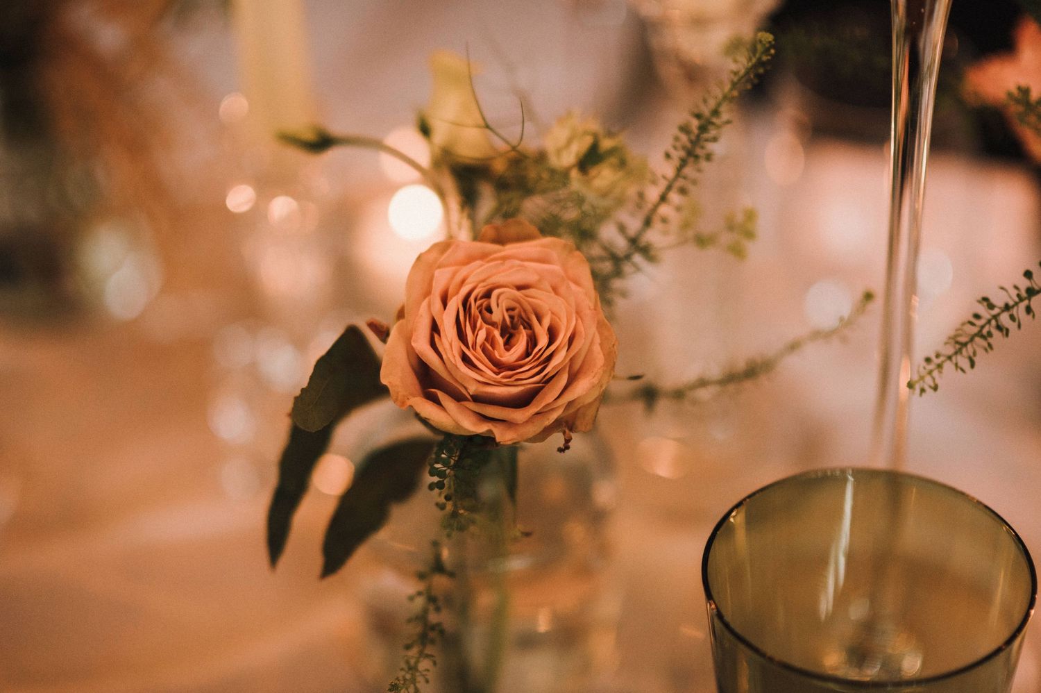 Peachy-pink rose with delicate foliage in a glass vase at a dinner setting.