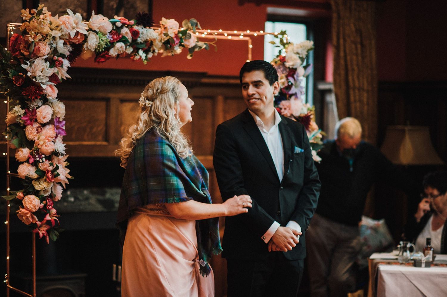 Couple standing by floral-decorated fireplace during wedding reception.